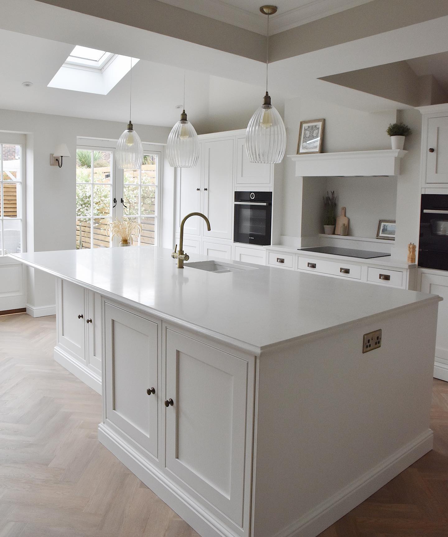 Soft white shaker cabinets with large island, three ribbed glass pendant lights, aged brass cup pulls, built-in Bosch oven, and herringbone wood floor