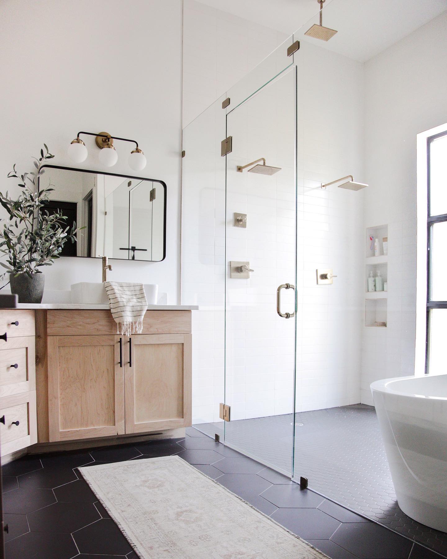 Bright white farmhouse bathroom with light wood shaker vanity, matte black hexagon floor tile, double rainfall shower behind frameless glass, freestanding tub by a black framed window, and olive branch arrangement