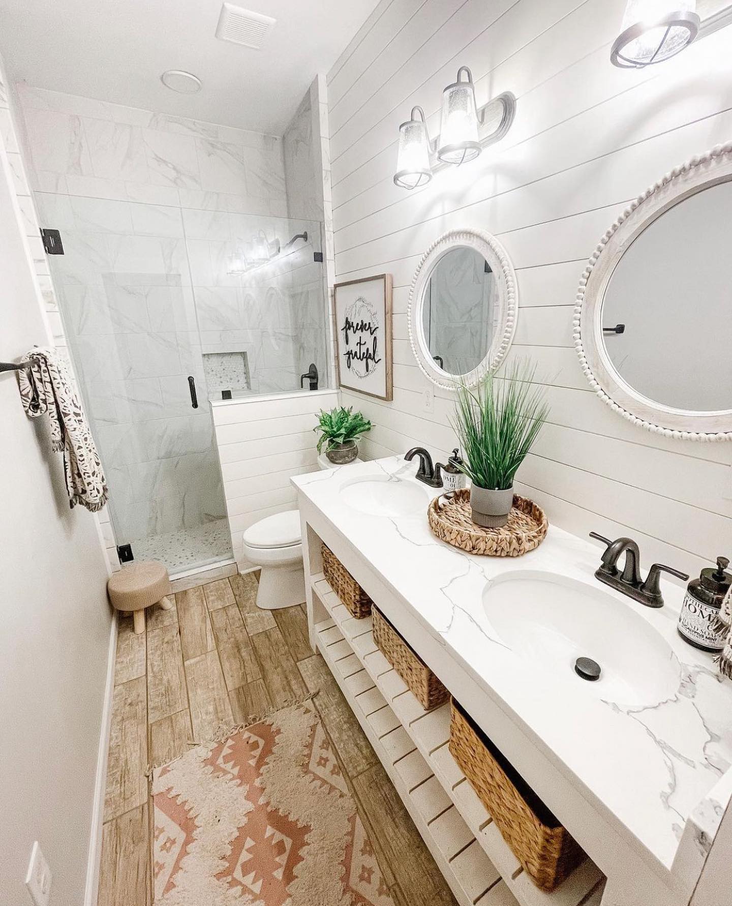 White shiplap farmhouse bathroom with double white vanity, marble countertop, two beaded round mirrors, woven baskets on open shelf, wood-look tile floor, pink Aztec rug, and marble tiled walk-in shower