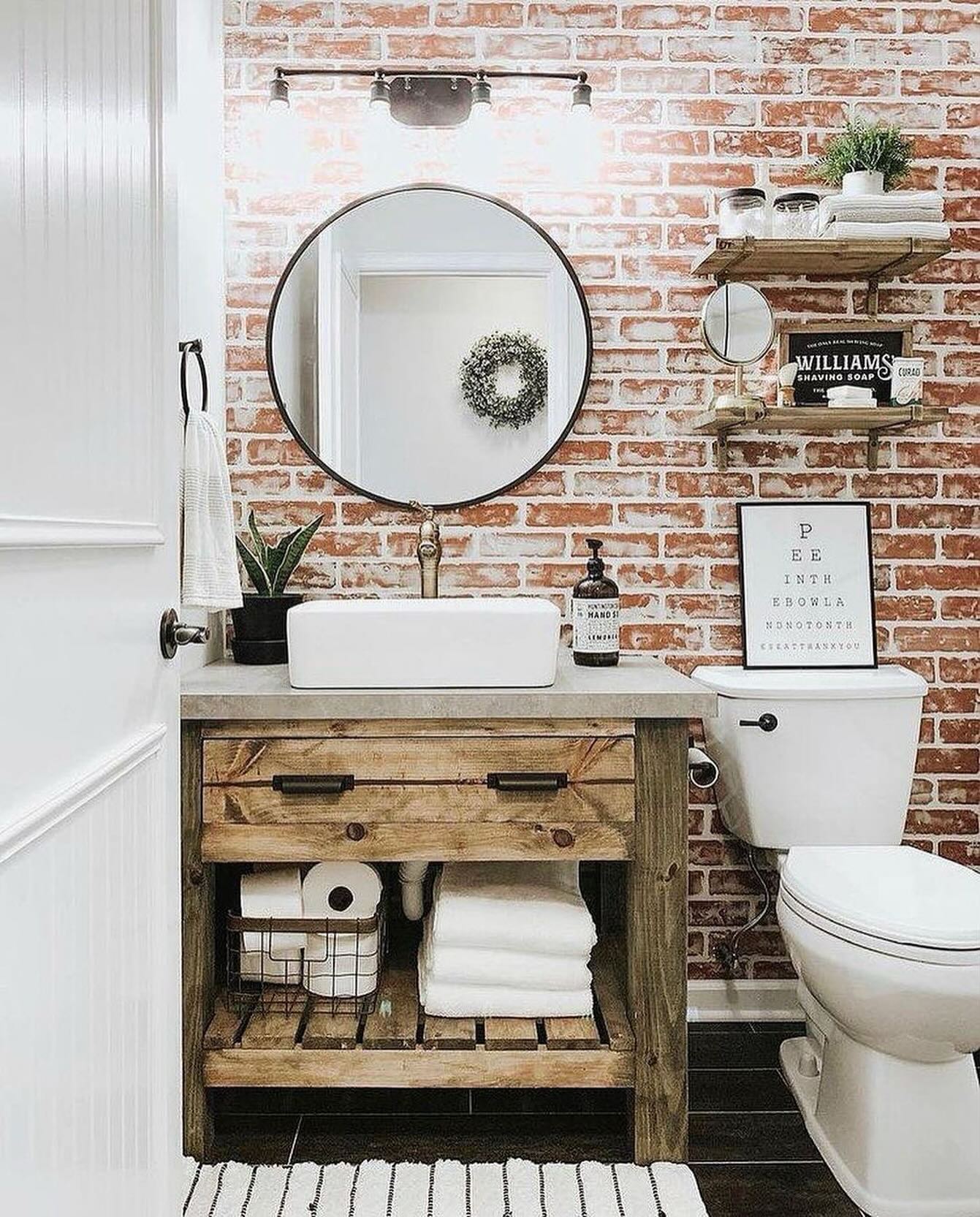 Farmhouse powder room with full red brick accent wall, round black framed mirror, rustic wood vanity with concrete countertop and white vessel sink, vintage Williams shaving soap sign on open wood shelves, snake plant, and striped black and white runner