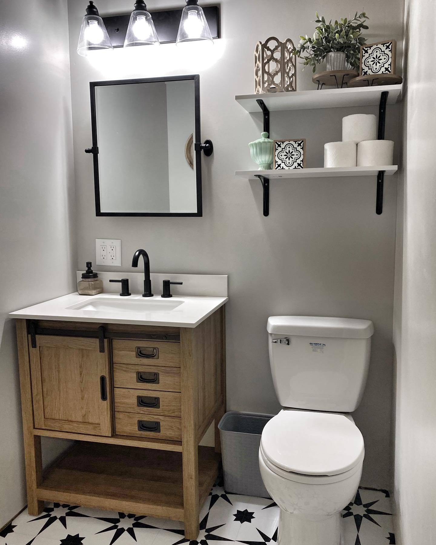 Small farmhouse bathroom with warm wood vanity featuring a sliding mini barn door, white quartz top, matte black single handle faucet, black pivot mirror, three-light vanity bar, black and white star pattern cement floor tile, and floating white shelves above the toilet