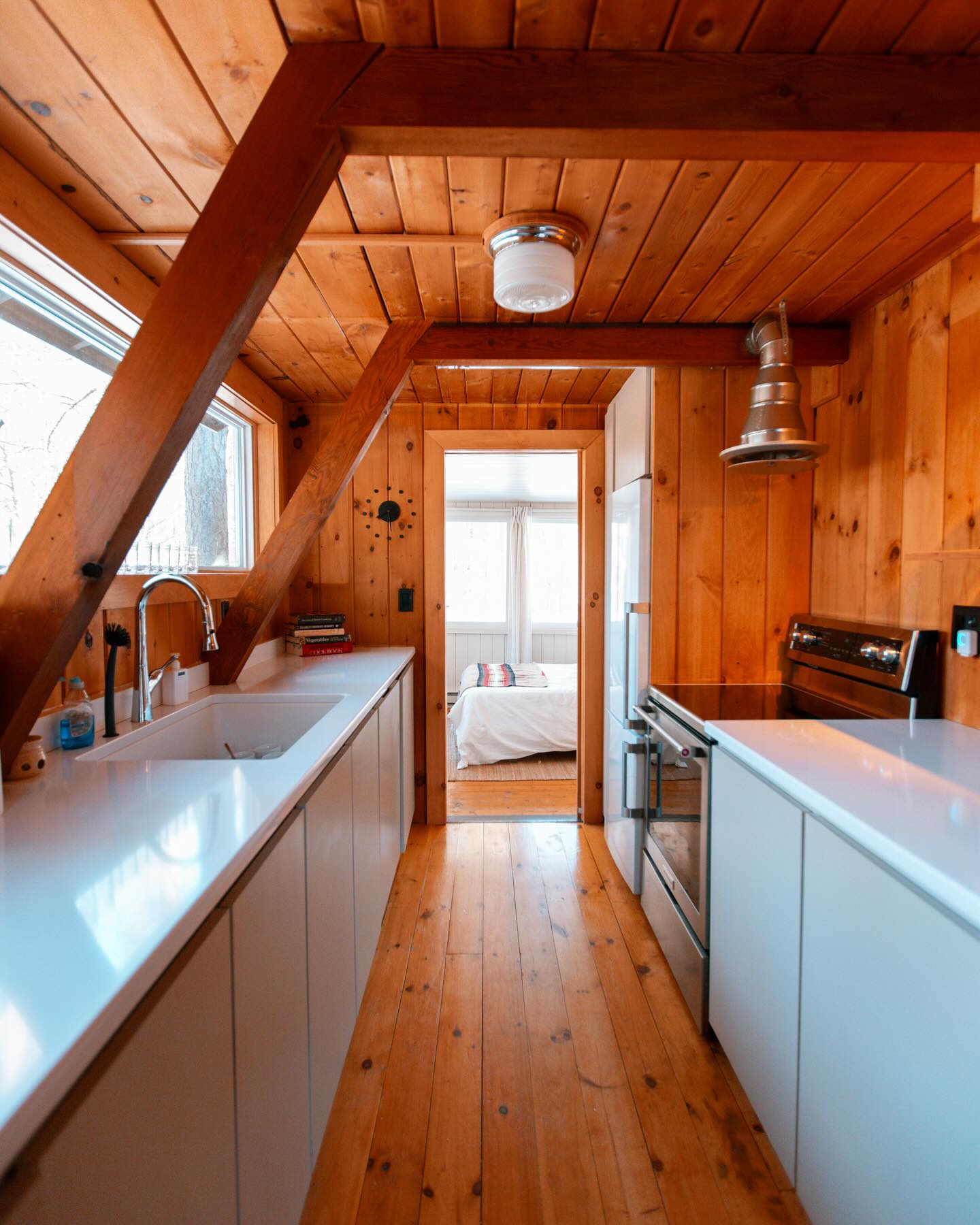 Narrow A-frame cabin kitchen with warm pine plank walls and ceiling, exposed diagonal beam, white lacquered base cabinets, bright white countertops, and a view through to a bedroom at the far end