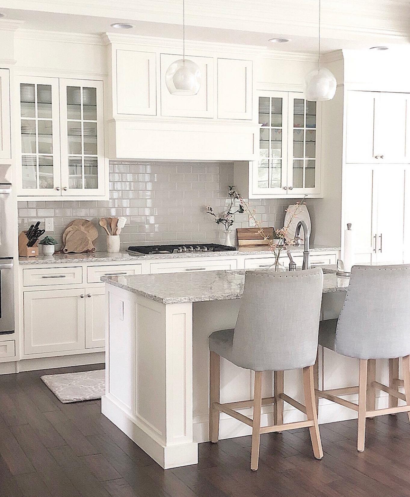All-white shaker kitchen with glass front upper cabinets flanking a tall paneled range hood, two gray upholstered counter stools with light wood legs, light gray subway tile backsplash, speckled granite island counter, and dark hardwood floor