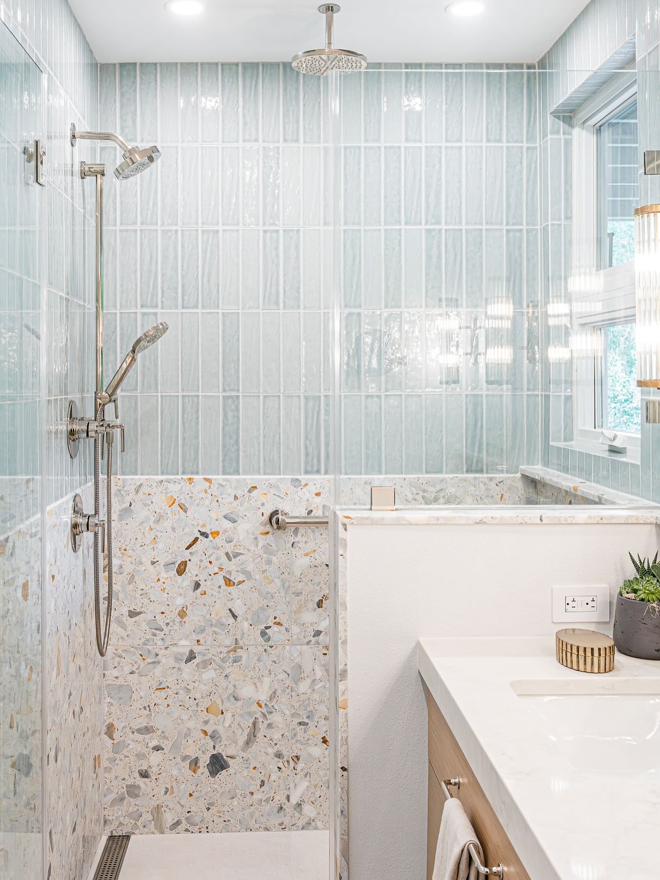 Walk-in shower with pale aqua vertical stacked glass tiles on the upper walls, a chunky multi-color terrazzo panel on the lower wall, polished nickel fixtures, and a marble topped vanity in the foreground
