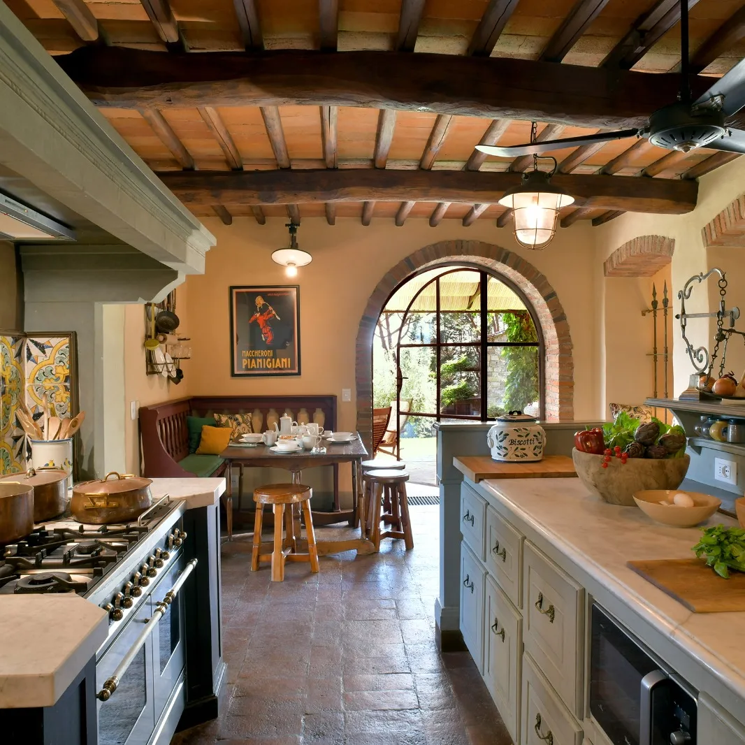 Rustic Tuscan villa kitchen with exposed terracotta tile ceiling and dark wood beams, arched brick-trimmed window opening to garden, copper pots on a dark pro range, vintage Macheroni Pianigiani poster, hand-painted ceramic tile panel beside the range, and terracotta tile floor