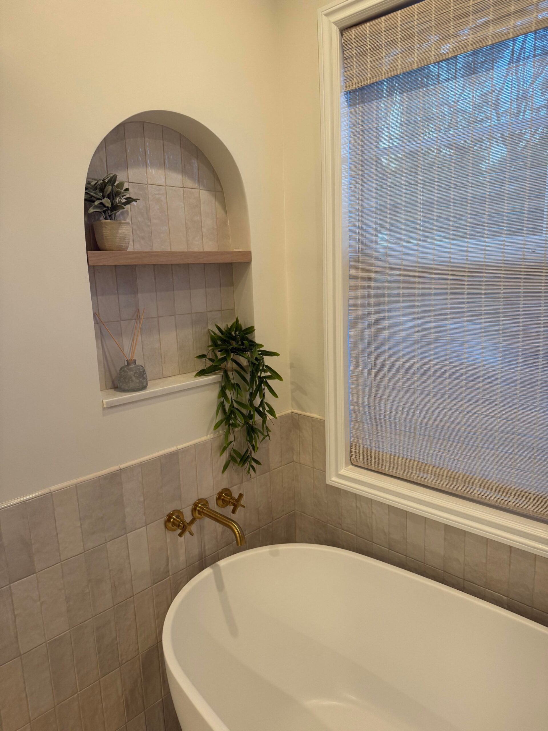 Freestanding tub corner with wall-mount brass tub filler, soft gray square tile wainscoting, and an arched built-in niche with a wood shelf, trailing plant, and small reed diffuser.