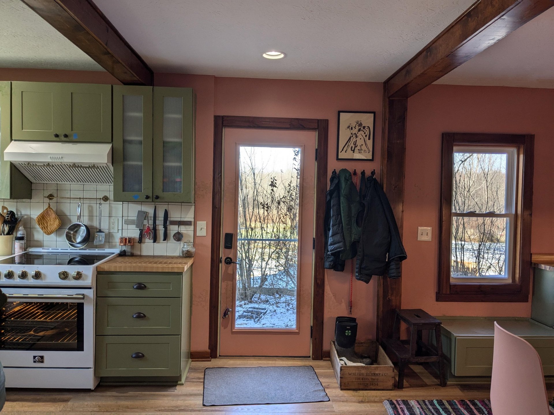 View toward the back glass door with coral pink walls, dark stained wood door frames, a white Forno range on the left, sage green cabinets, and a wooden crate with muck boots on the floor.