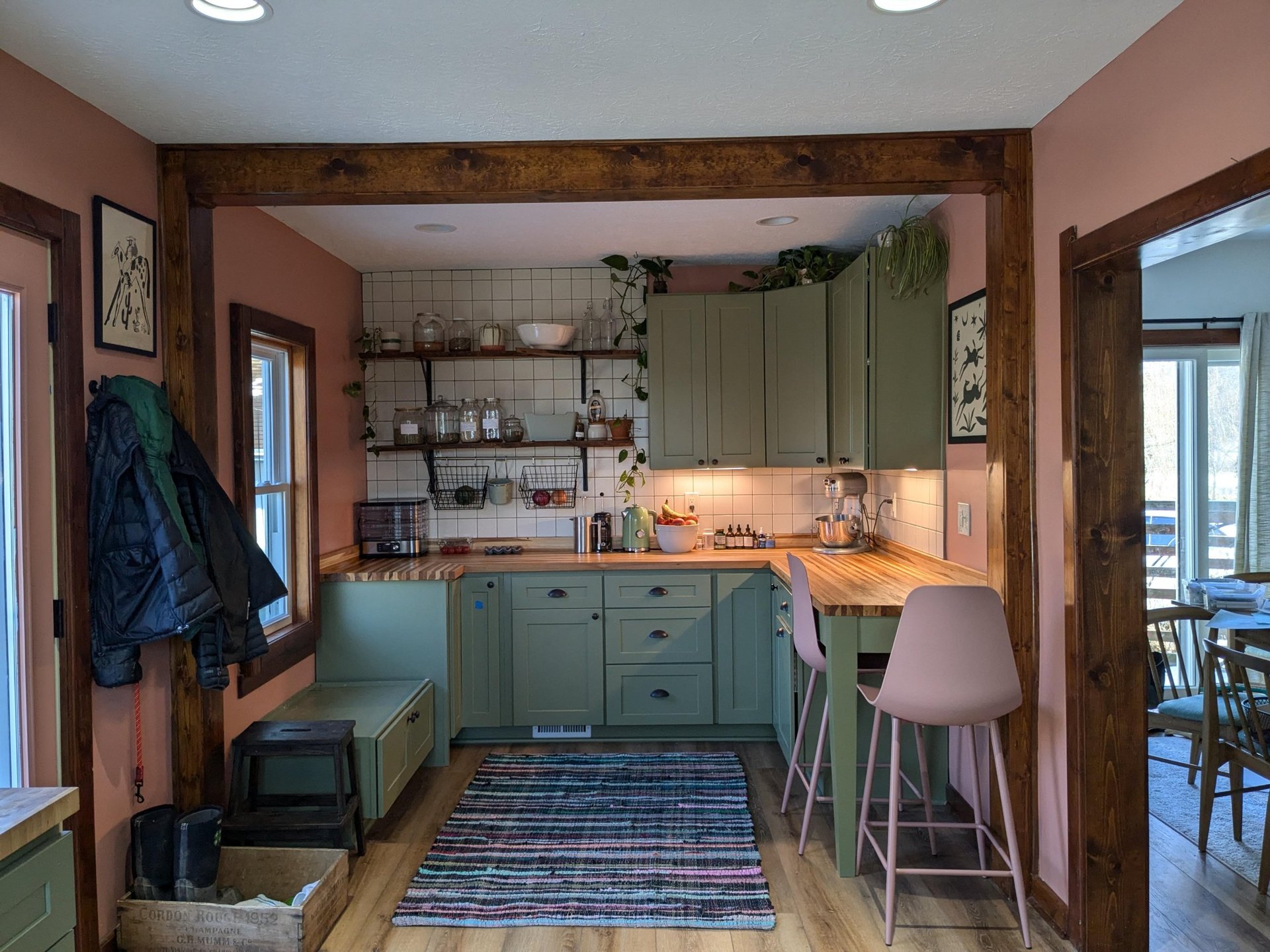 Finished back wall with sage green lower cabinets, butcher block counter with a KitchenAid mixer and herbal jars, open live-edge wood shelves made from trees on the property, a pink striped runner, and two pink barstools at a peninsula.