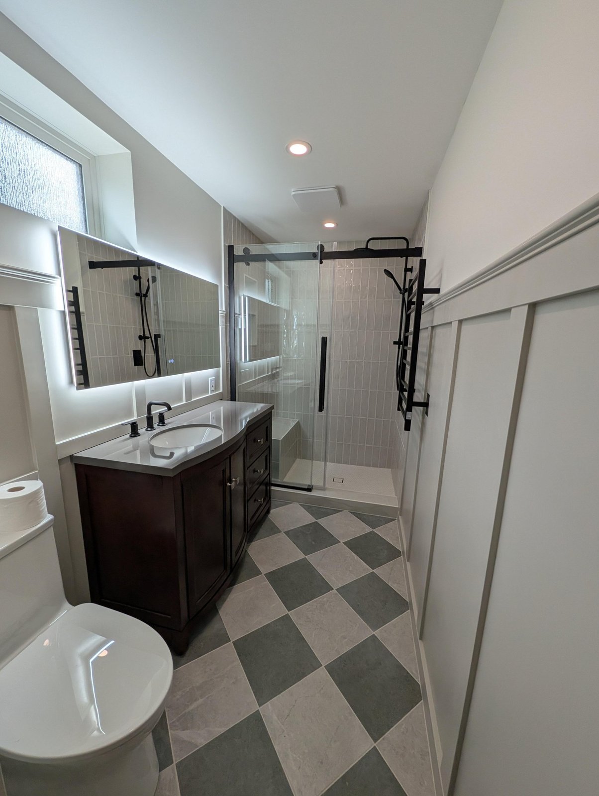 Dark wood single-sink vanity with a gray quartz top, matte black faucet, a glowing backlit LED mirror above it, and the glass shower reflected on the right.