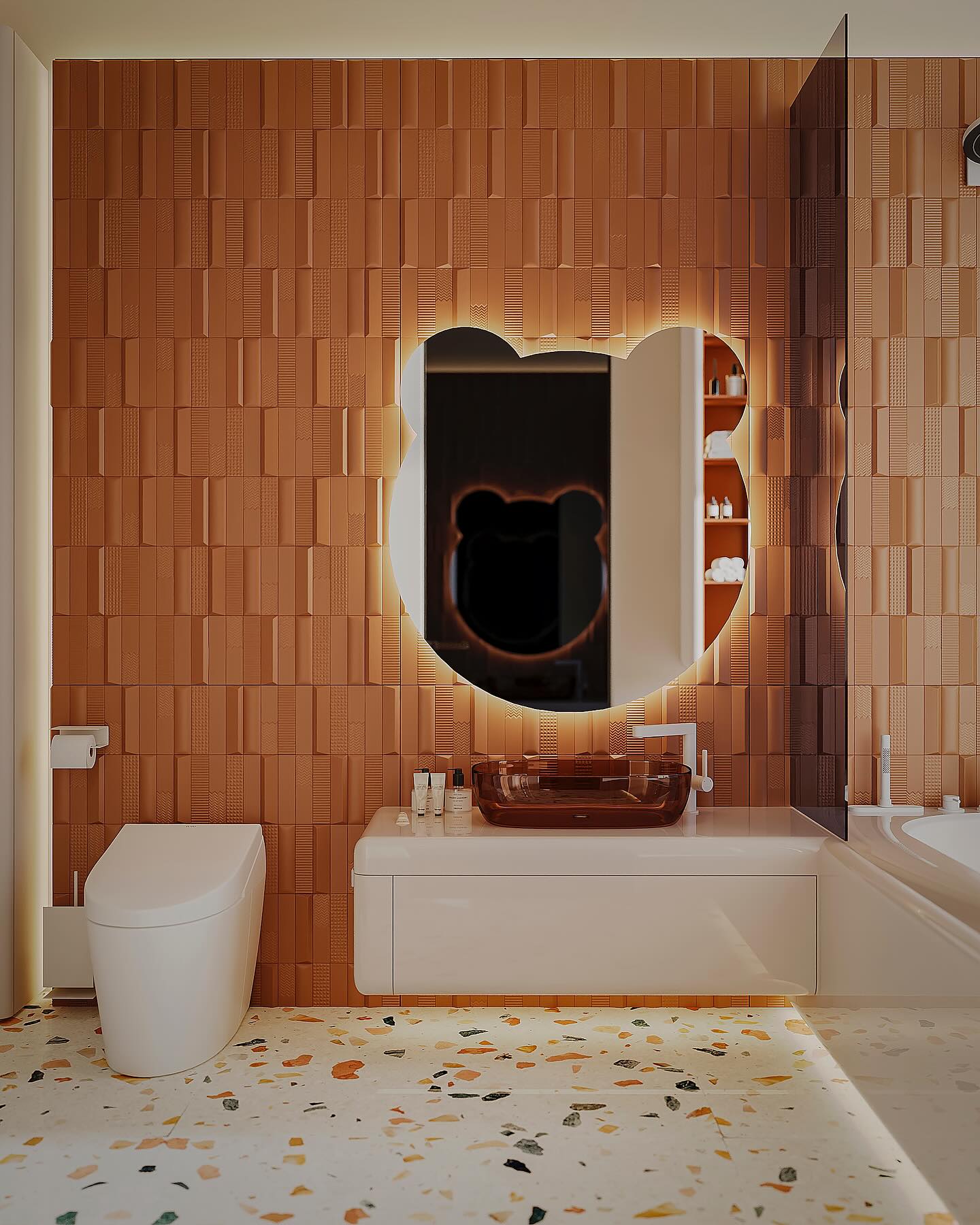 Bear-shaped backlit mirror on a terracotta brick-textured tile wall above a floating white vanity with amber vessel sink and confetti terrazzo floor