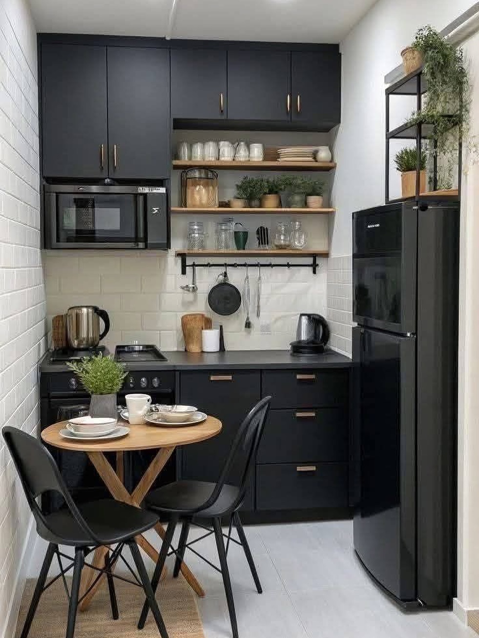 Small kitchen with floor to ceiling matte black cabinets, brass handles, white brick wall, open wood shelves styled with greenery, and a small round wood dining table with two black chairs