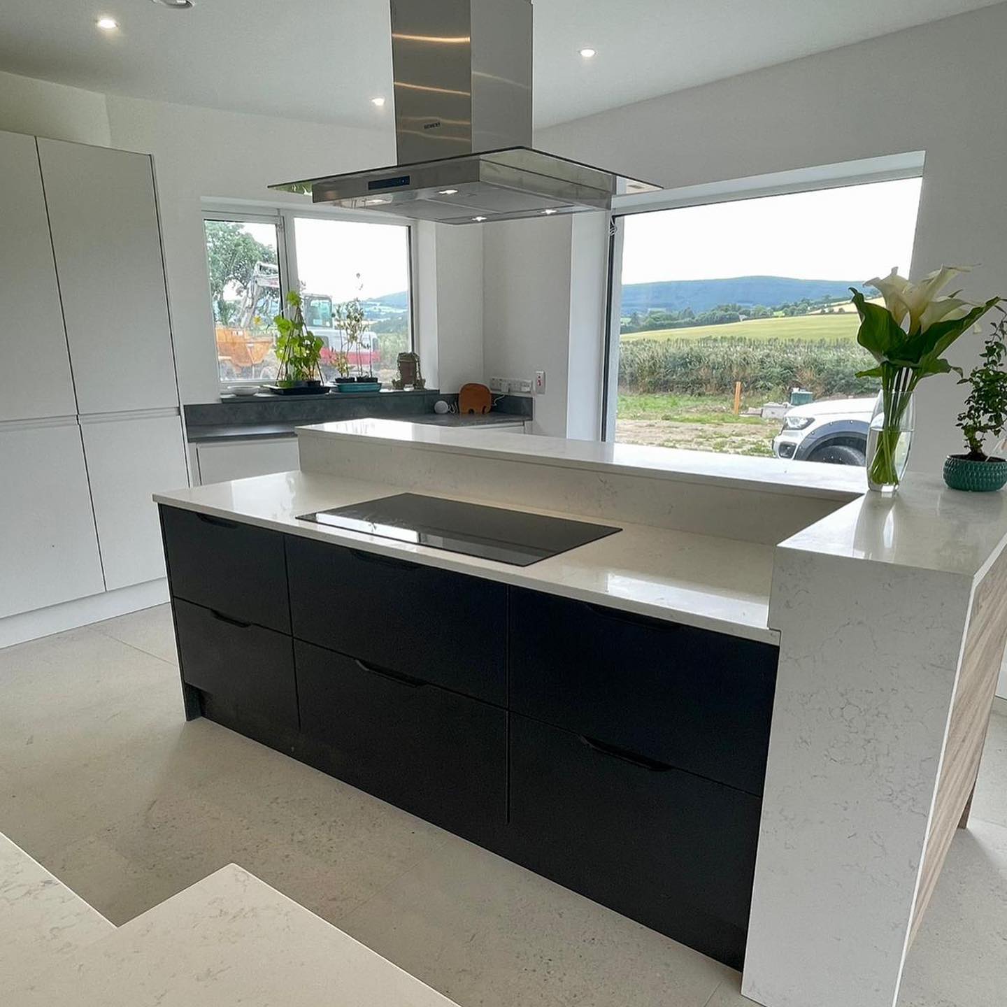 Black flat-front handleless island cabinets with white quartz waterfall countertop, stainless steel island hood, and a view of green hills through the window