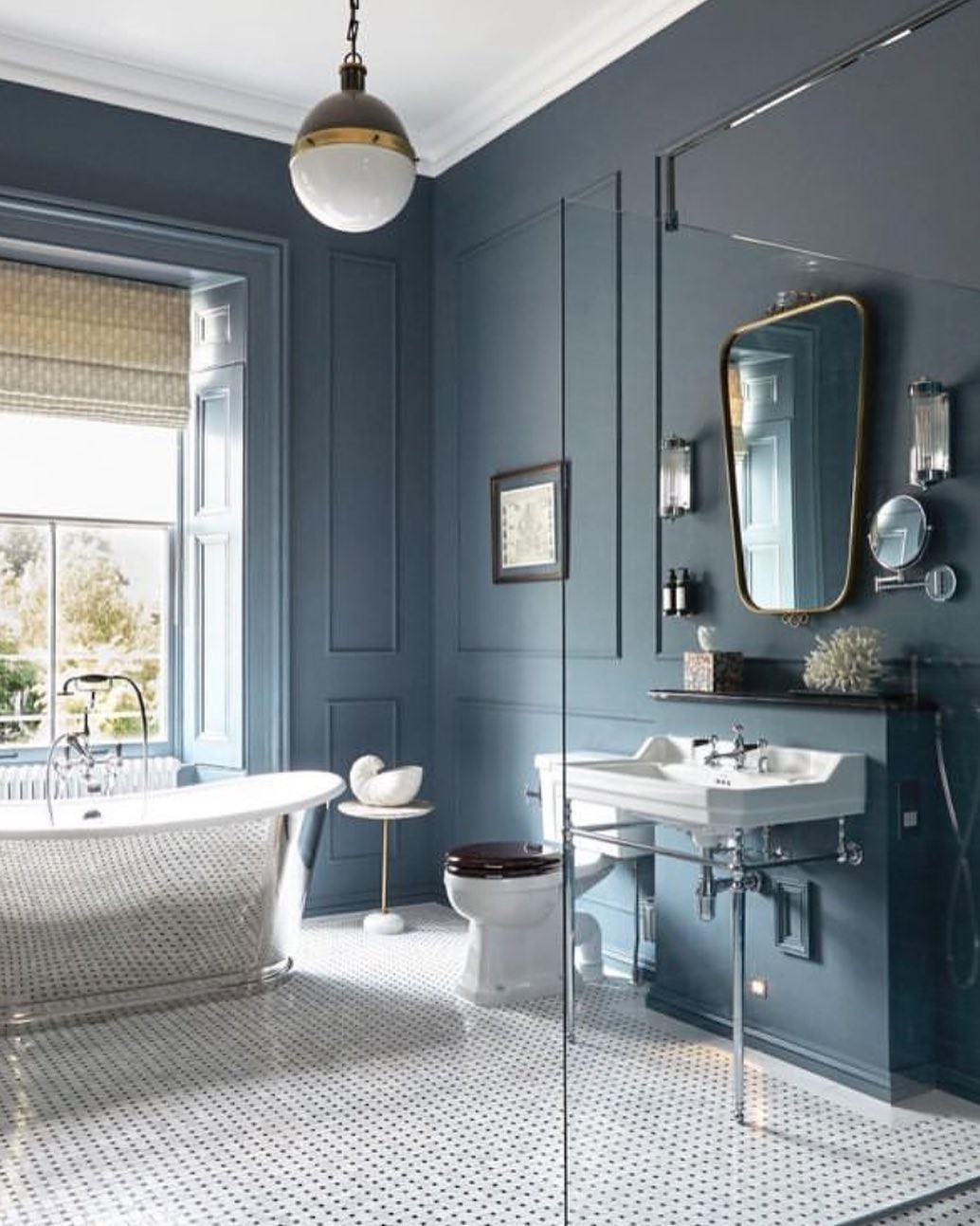 Blue-gray painted paneled bathroom walls with a white clawfoot tub by the window, white hexagon tile floor, pedestal sink with chrome legs, and gold-framed arched mirror