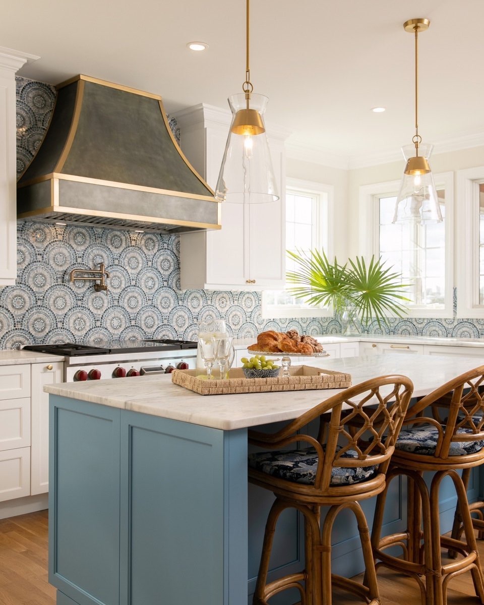 Coastal kitchen with a blue circular mosaic tile backsplash behind the range, a dark pewter hood with gold trim bands, clear glass pendants with brass tops, white cabinets, a soft blue island with marble top, and two rattan bar chairs