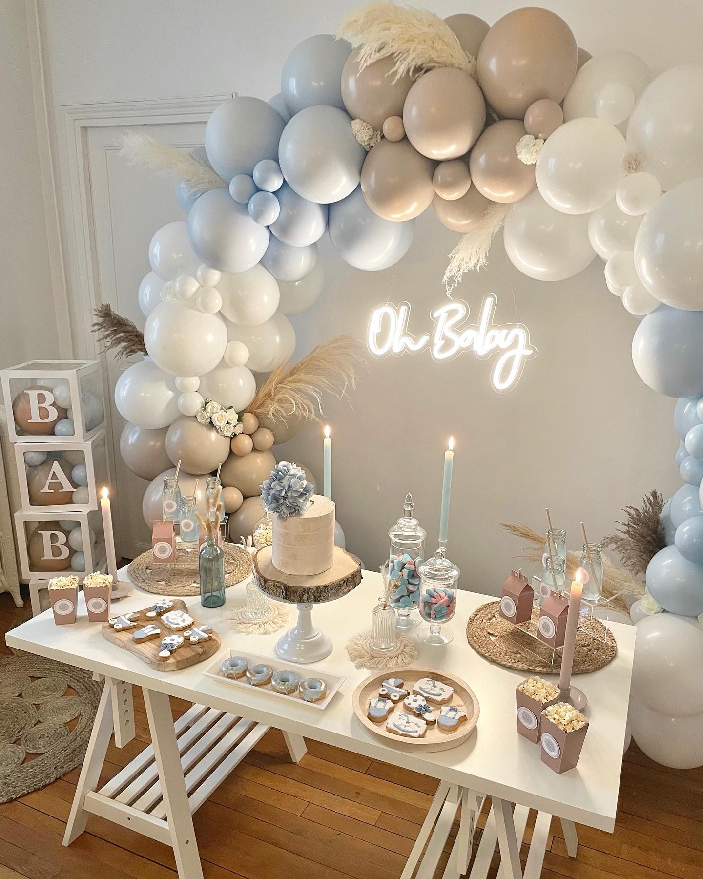 Dusty blue, tan, and white balloon arch backdrop with pampas grass, oh baby neon sign, BAB blocks, and a small wood slice cake on a white table
