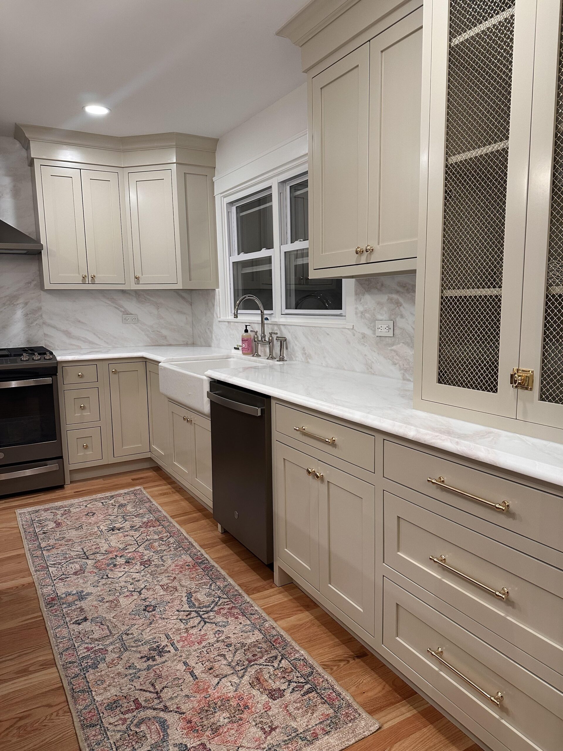 Taupe shaker cabinets with warm brass bar pulls and knobs, a white apron farmhouse sink, polished nickel bridge faucet, and a brass mesh cabinet insert on the right.