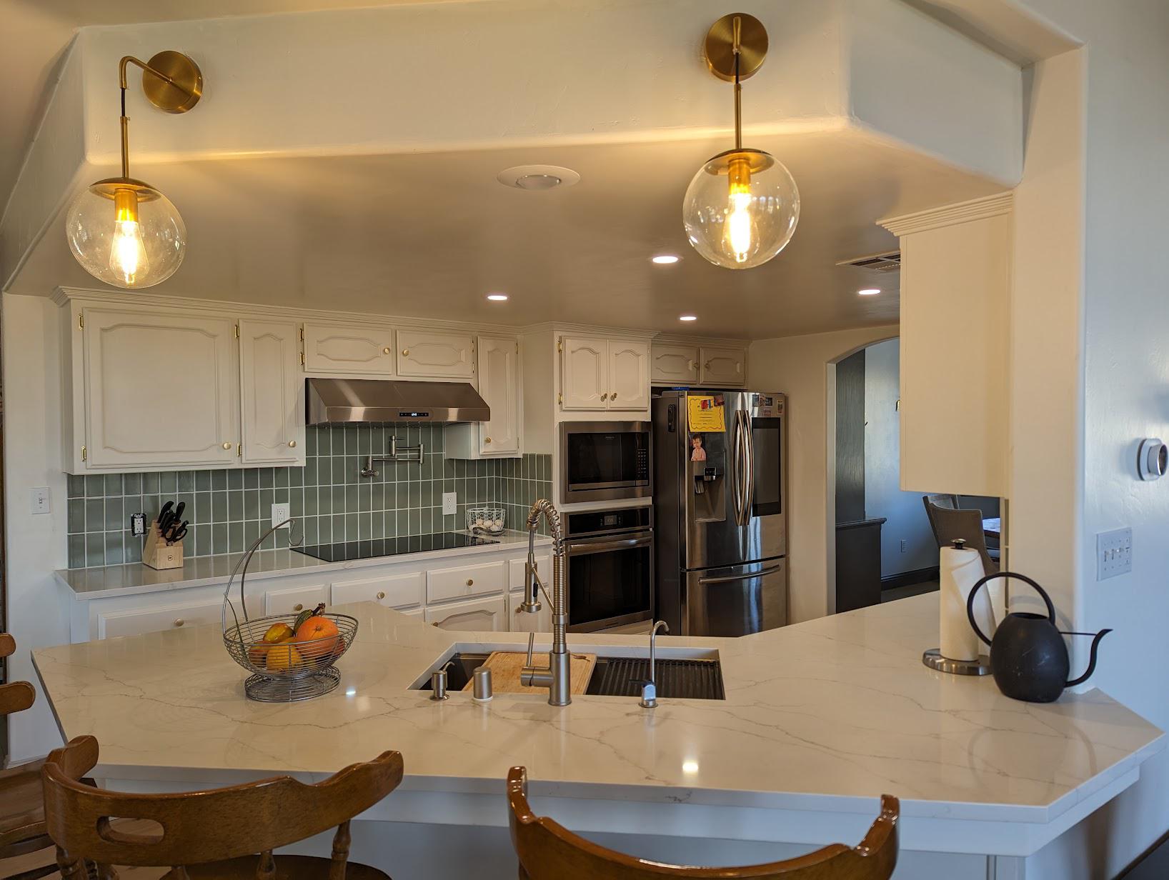 View across a white quartz peninsula with soft gray veining toward the renovated kitchen, two brass wall sconces with clear globe shades on either side of the soffit, sage green backsplash, induction cooktop, and stainless fridge in the back.