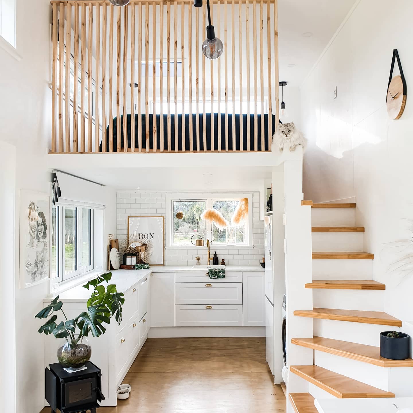 Bright white tiny house kitchen with a sleeping loft above, wood slat railing, subway tile backsplash, brass hardware on white Shaker cabinets, and a wooden staircase on the right