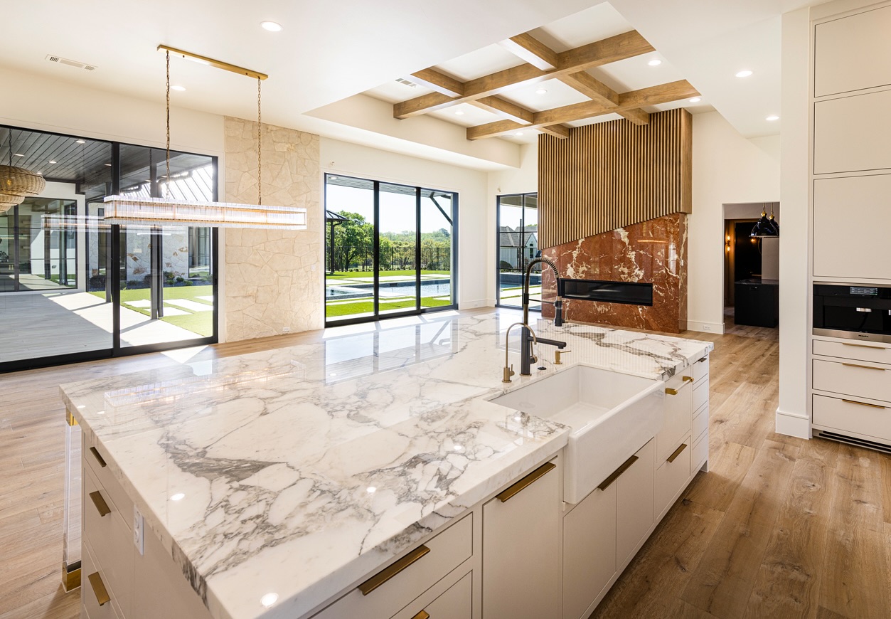 Large white kitchen with dramatic Calacatta marble island, exposed wood beam ceiling, white farmhouse sink, and open view through sliding doors to a pool