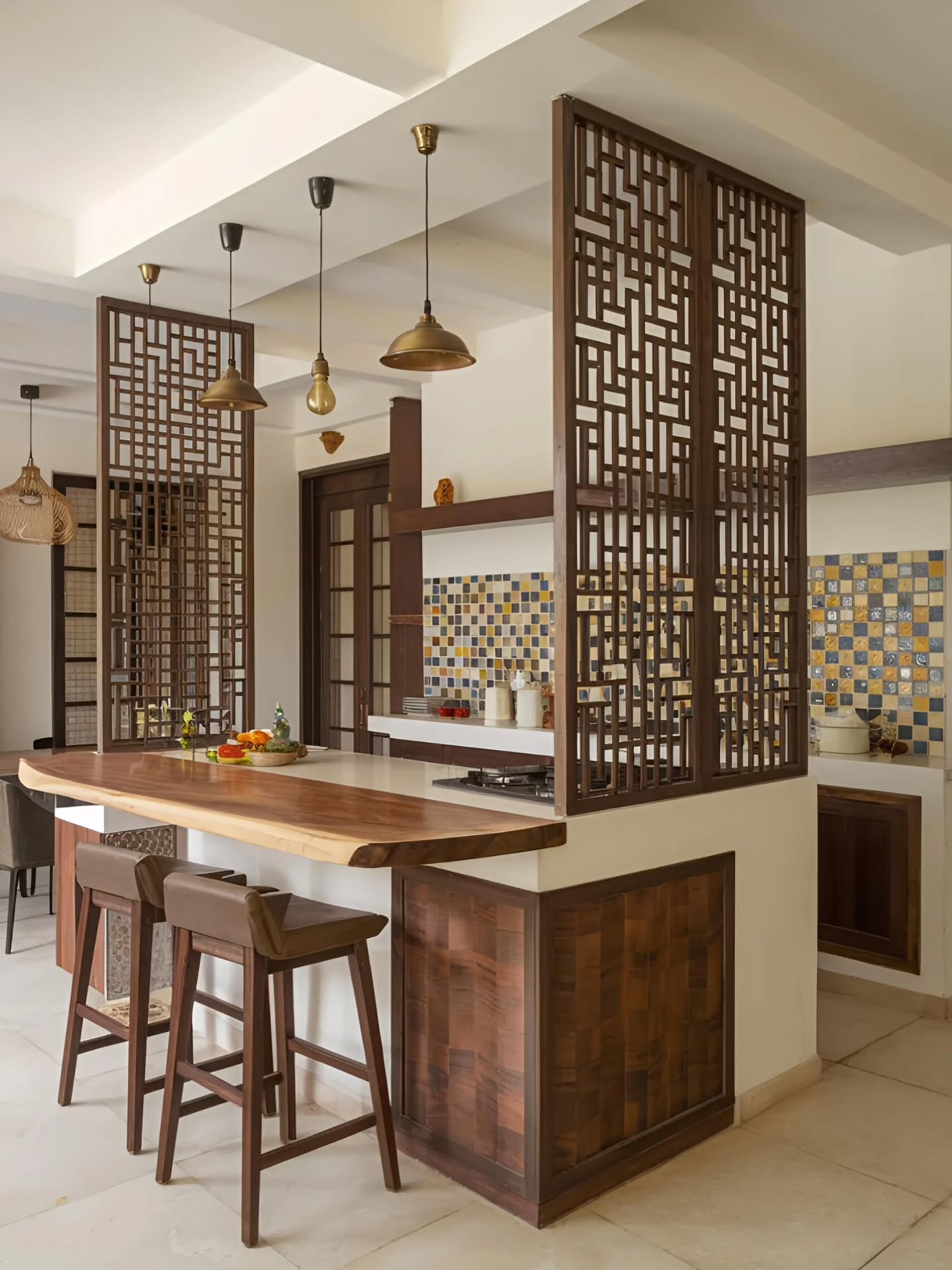 Open kitchen seen through dark wood carved geometric fretwork screens, with mixed yellow and blue mosaic tile backsplash, brass dome pendant lights with exposed Edison bulbs, a live-edge natural wood bar top, and brown leather bar stools with wood frames