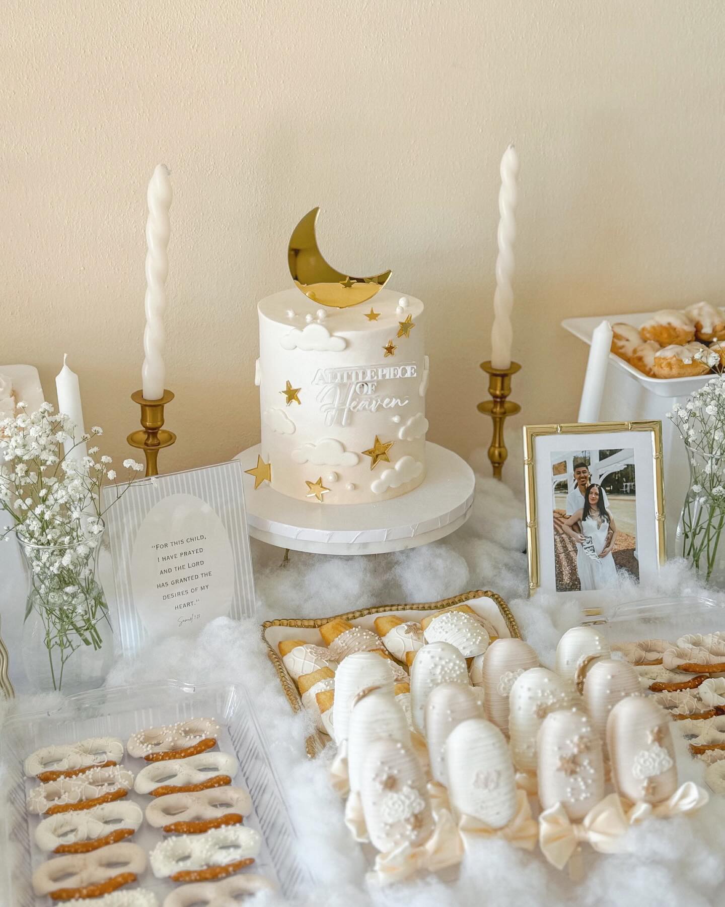 Celestial baby shower dessert table with a white cake topped with a gold moon, cloud fondant details, candles in brass holders, a Samuel scripture card, and white chocolate pretzel treats