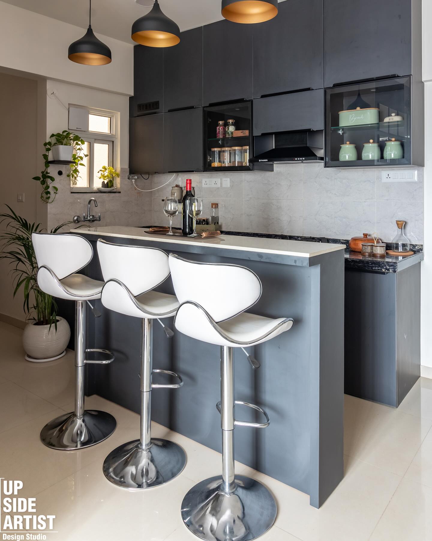 Open kitchen with dark charcoal cabinets, glass fronted upper storage holding mint canisters, three chrome and white bar stools, and a bottle of wine on the counter