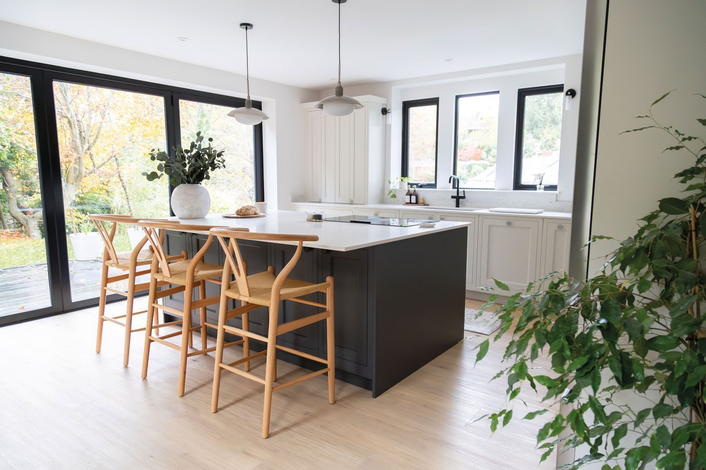 Charcoal gray minimalist kitchen island with light wood wishbone bar stools, white quartz countertop, and large sliding glass doors to a garden