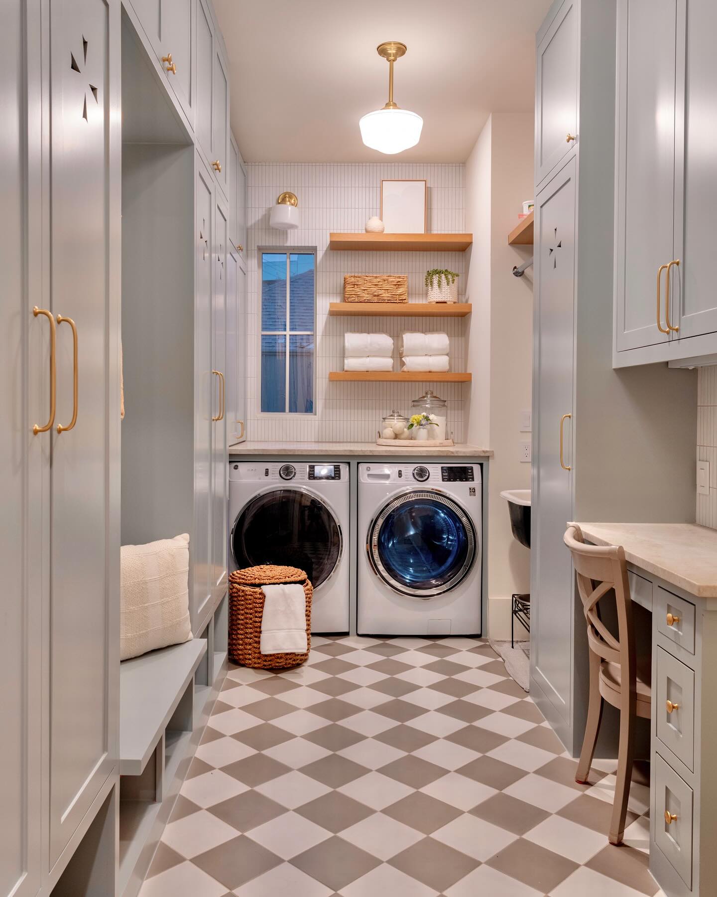 Light blue mudroom laundry room with floor-to-ceiling built-in cabinets with brass hardware, open wood shelves above a side-by-side washer dryer, a built-in desk with chair, and a tan and cream checkerboard tile floor