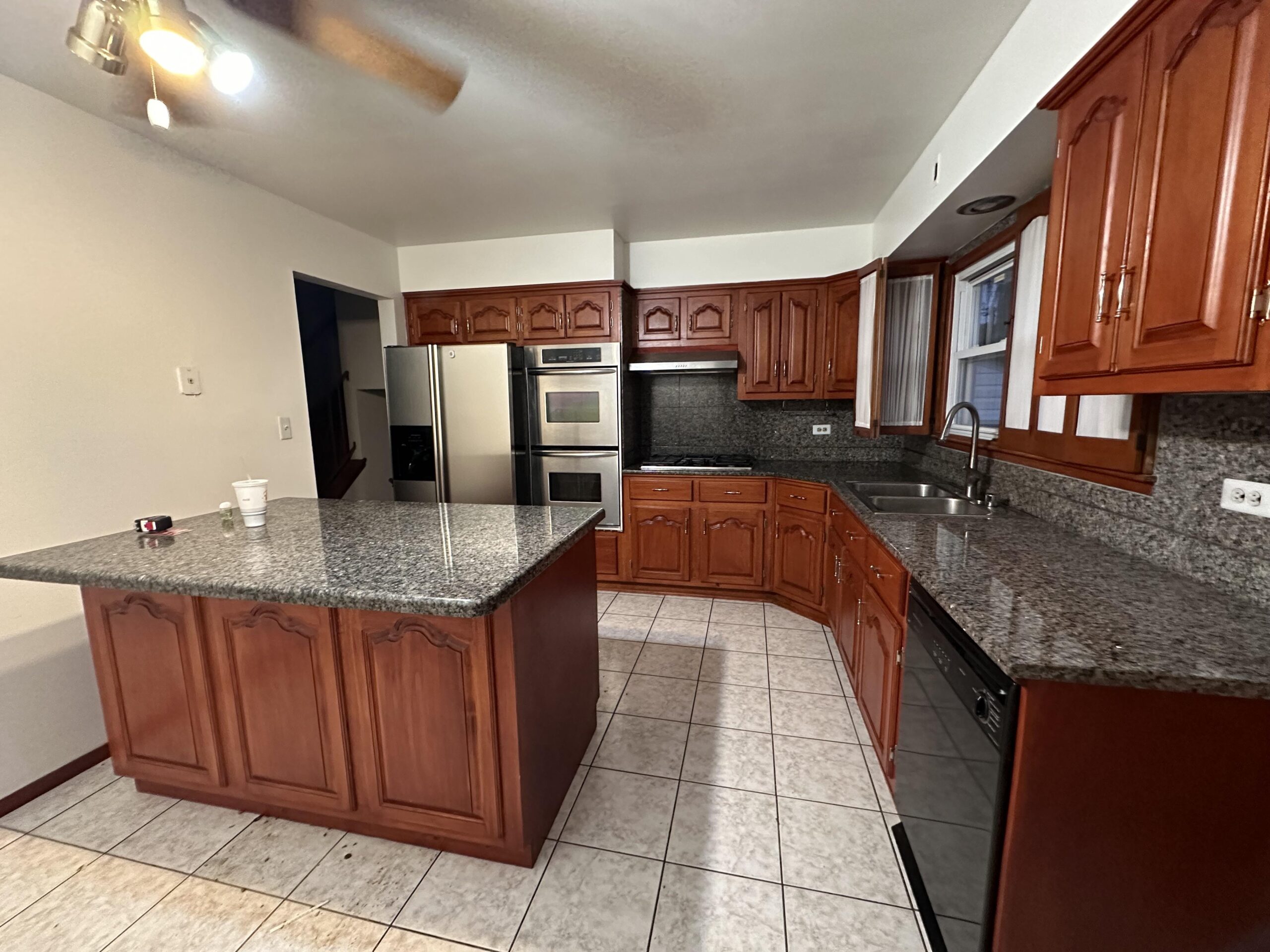 Wide shot of a U-shaped kitchen with cherry raised-panel cabinets, dark granite counters, ceiling fan, tile floor, and a bulky cabinet island in the middle.