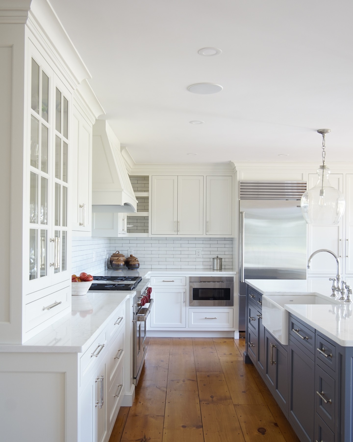 Classic white kitchen with glass front upper cabinets, raised panel white range hood, deep charcoal gray painted island with white quartz top and farmhouse sink, white subway tile backsplash, chrome bar pulls, and wide plank warm pine floors