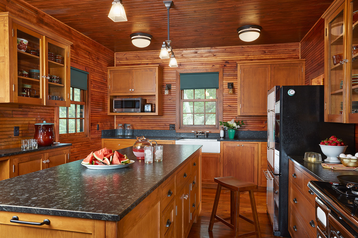 Classic wood cabin kitchen with horizontal pine plank walls and beadboard ceiling, dark granite countertops, glass-front wood cabinets, farmhouse sink, black fridge, and schoolhouse-style pendant lighting over a wood island