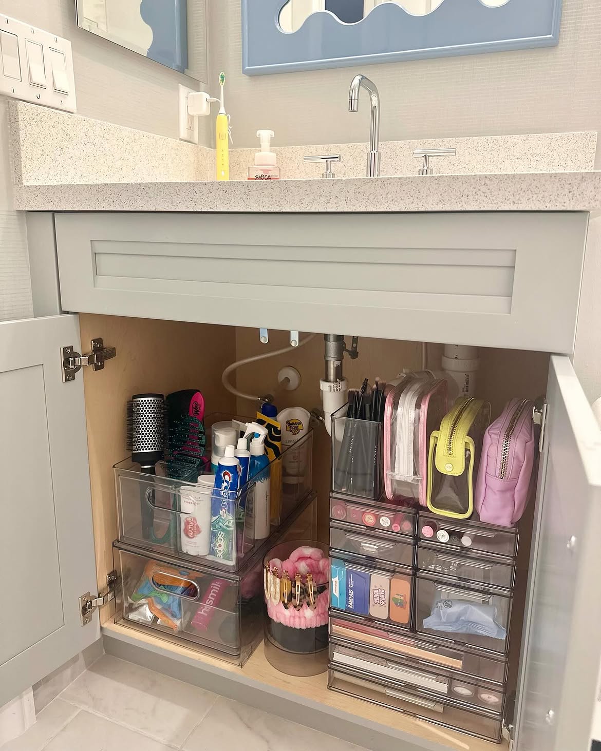 Open gray bathroom vanity under a half-door with clear stackable pull-out drawers on the left holding brushes and toiletries and a tall multi-drawer clear tower on the right holding makeup, bandaids, hair accessories and small cosmetic bags