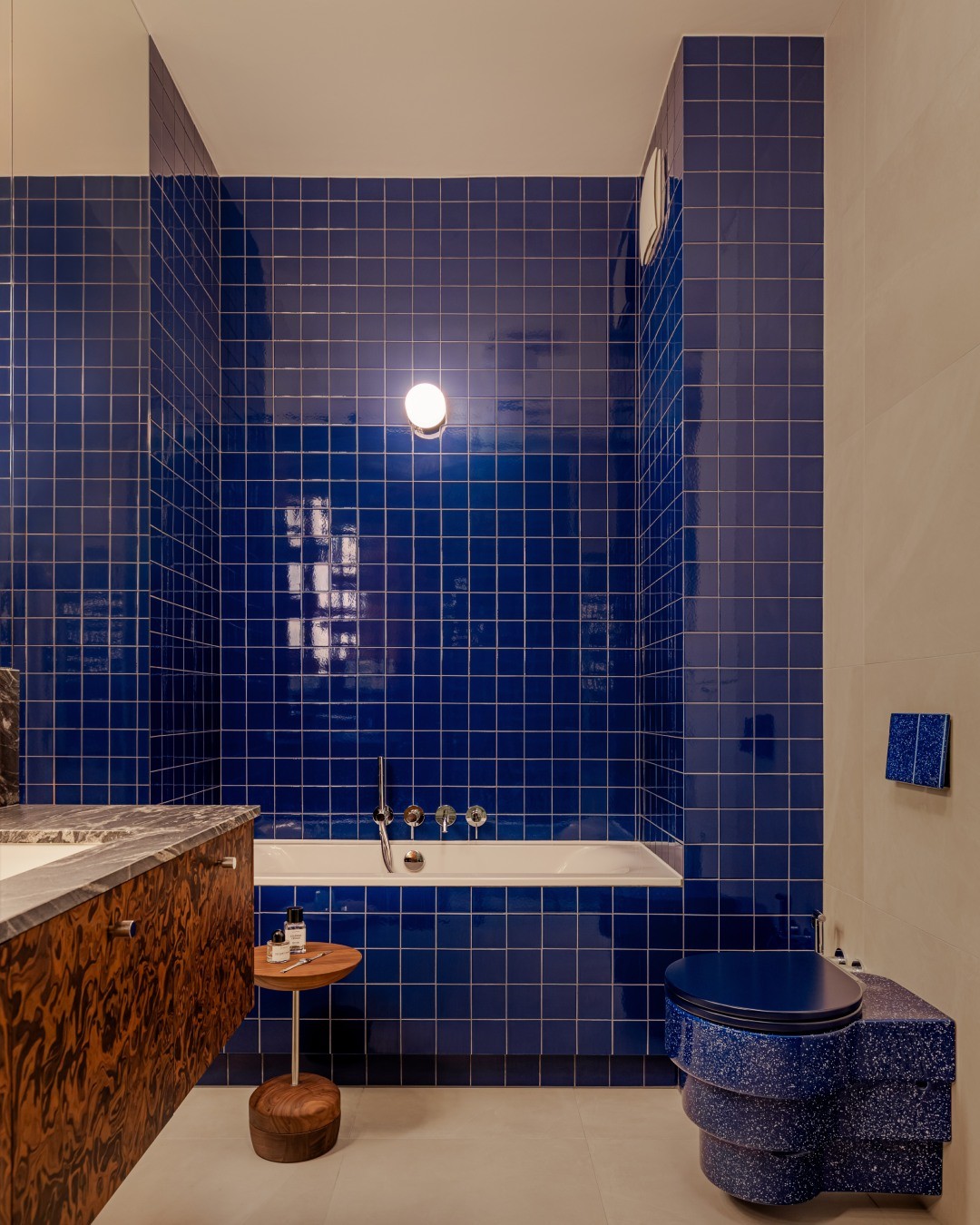 Cobalt blue square grid tile walls around a bathtub, with a burl wood vanity, gray marble counter, speckled cobalt blue wall-mounted toilet, and small walnut side table