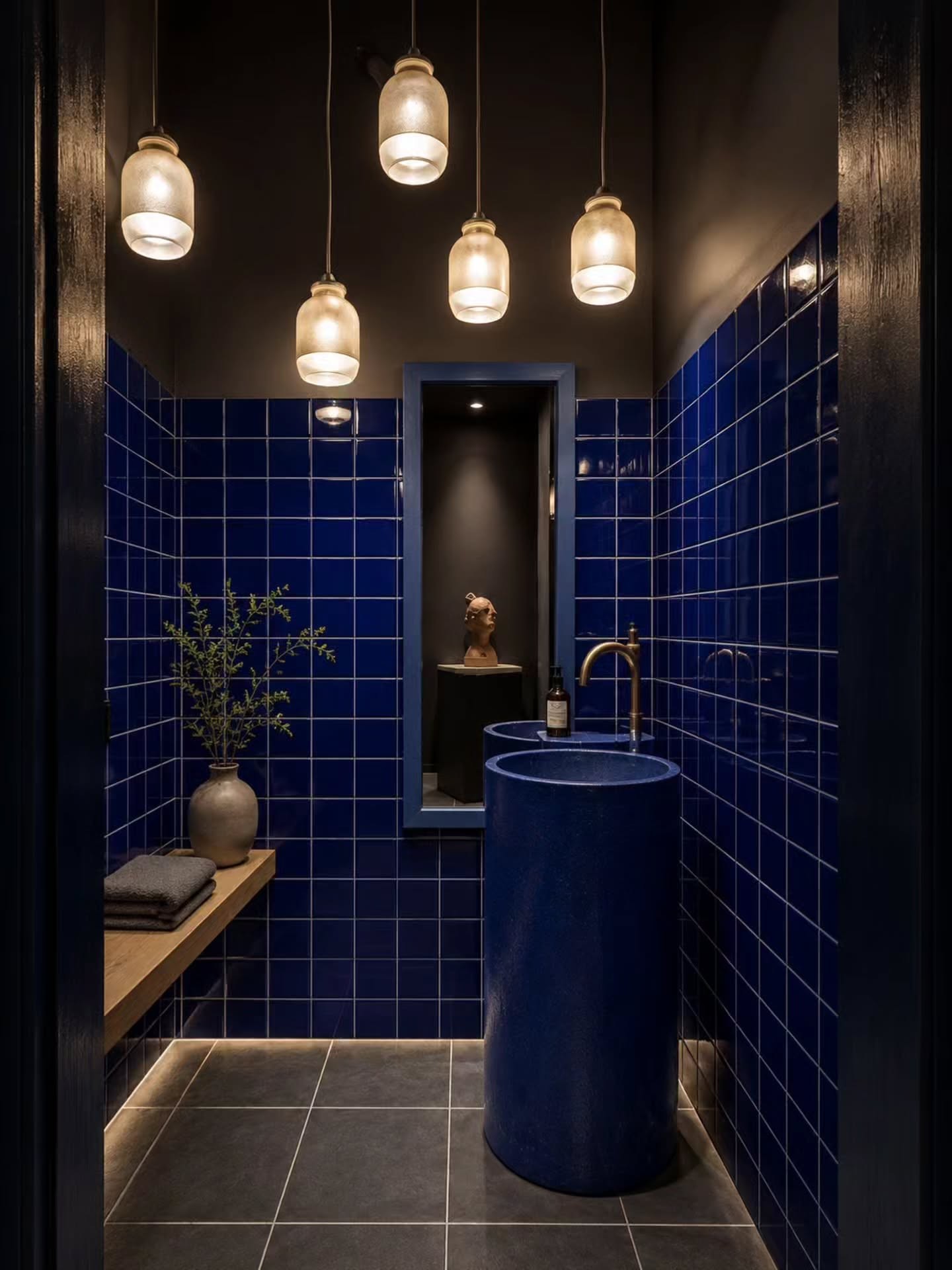 Dark cobalt blue square grid tile walls in a small powder room with a blue cylindrical pedestal sink, cluster of five amber glass pendant lights hanging from the ceiling, dark gray floor, and tall mirror