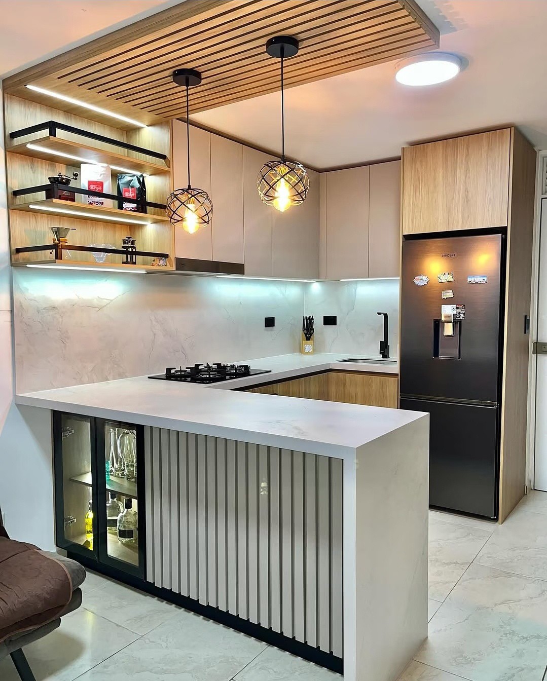 Compact open U-shaped kitchen with a slatted wood ceiling panel, white marble counters and backsplash, light wood and cream flat-panel cabinets, two black cage pendant lights with Edison bulbs, floating shelves with LED strip lighting, and a bar peninsula fronted with vertical gray slat paneling