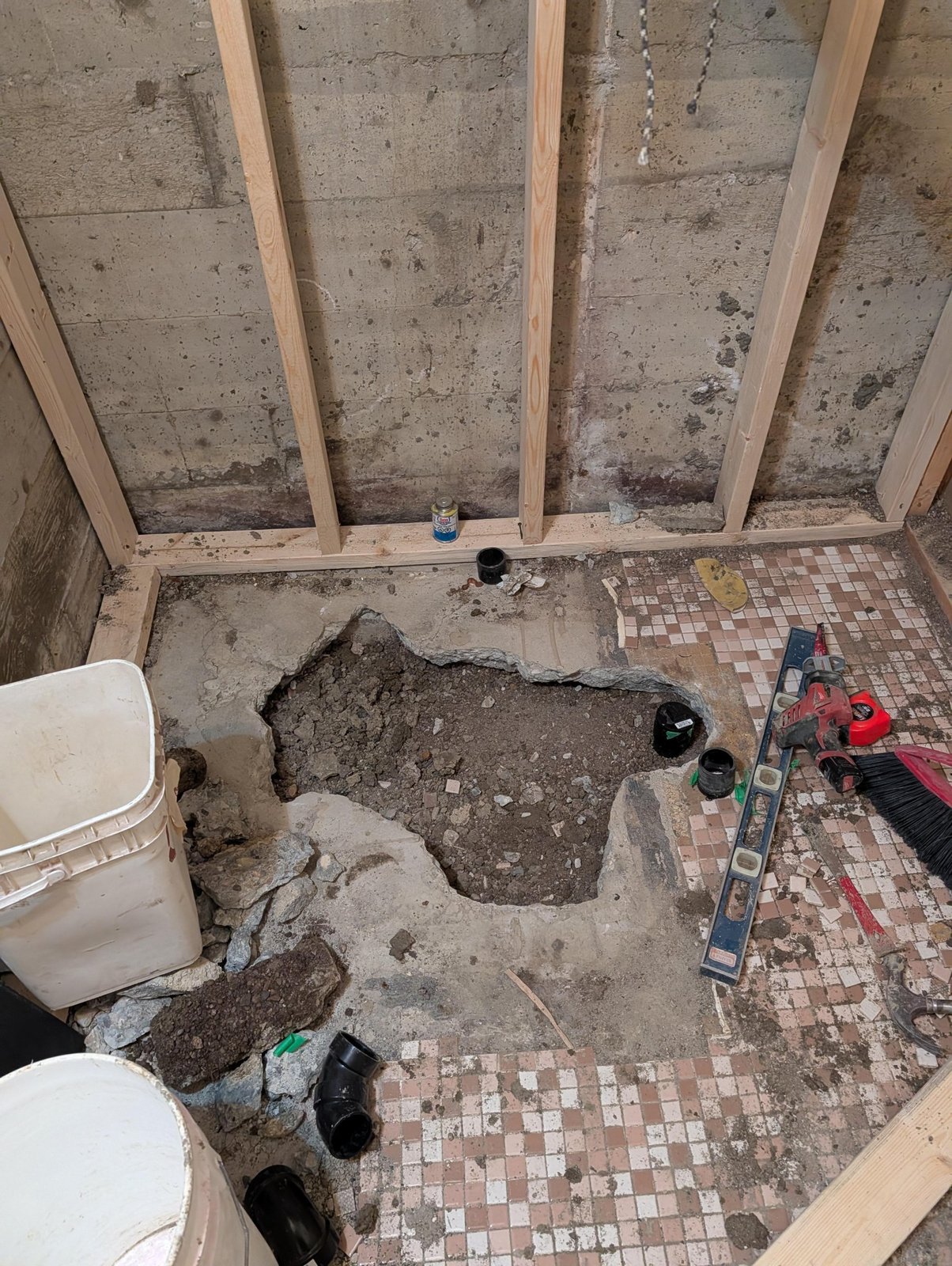 Bathroom floor with a jagged hole chopped through the concrete slab to expose dirt and black ABS drain fittings, with a level and tools lying beside it.