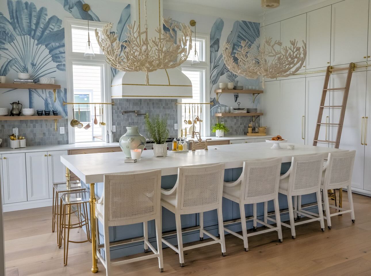 Coastal kitchen with two large white coral branch chandeliers hanging over a long marble island, soft blue palm leaf wallpaper mural on the walls, white cane barstools, white cabinets, and a white and gold range hood