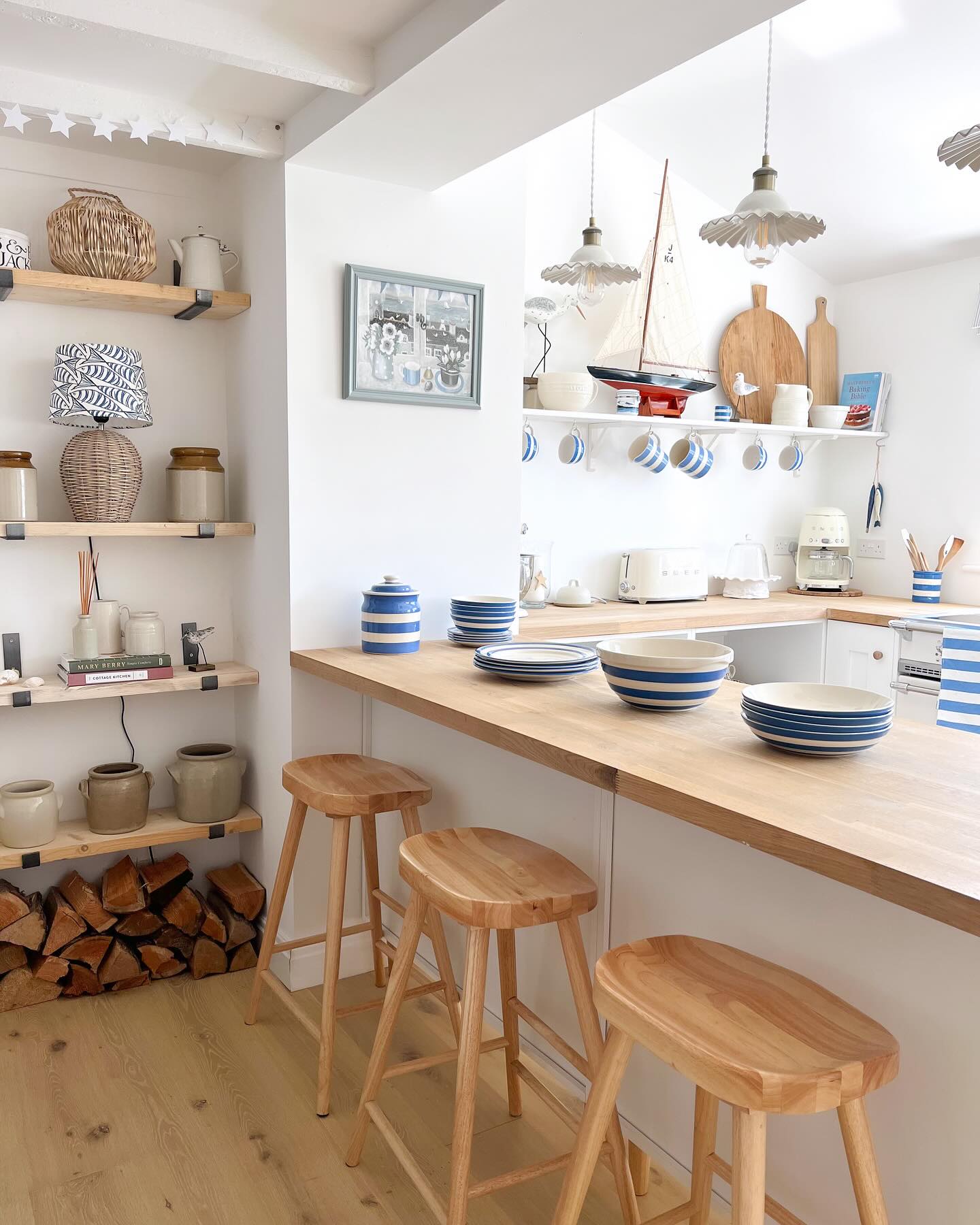 Small cottage coastal kitchen with butcher block wood counters, open wall shelves stacked with blue and white striped Cornish pottery bowls and jars, a small wooden model sailboat on the shelf, three round wood bar stools, and firewood stored underneath the counter