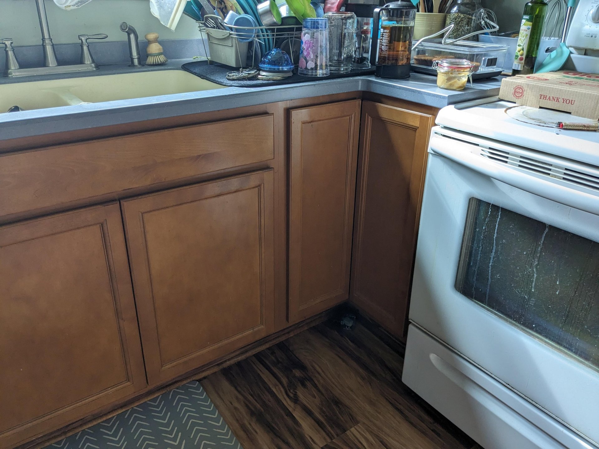 Close-up corner of the before kitchen with orange oak cabinets, a cluttered sink, a greasy white stove, and a pizza box on the stovetop.