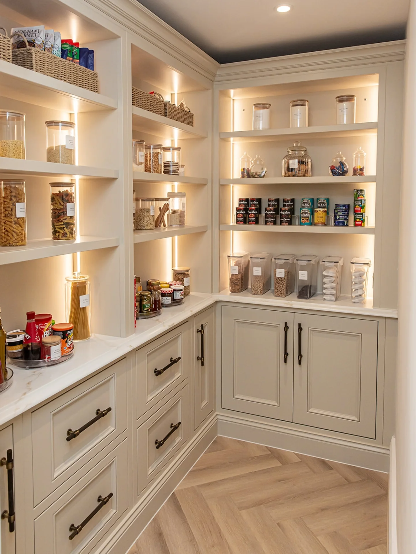 Cream Shaker-style pantry with floor-to-ceiling open shelves, warm under-shelf strip lighting, white marble counter, bronze pulls, dark painted ceiling, and herringbone wood floor