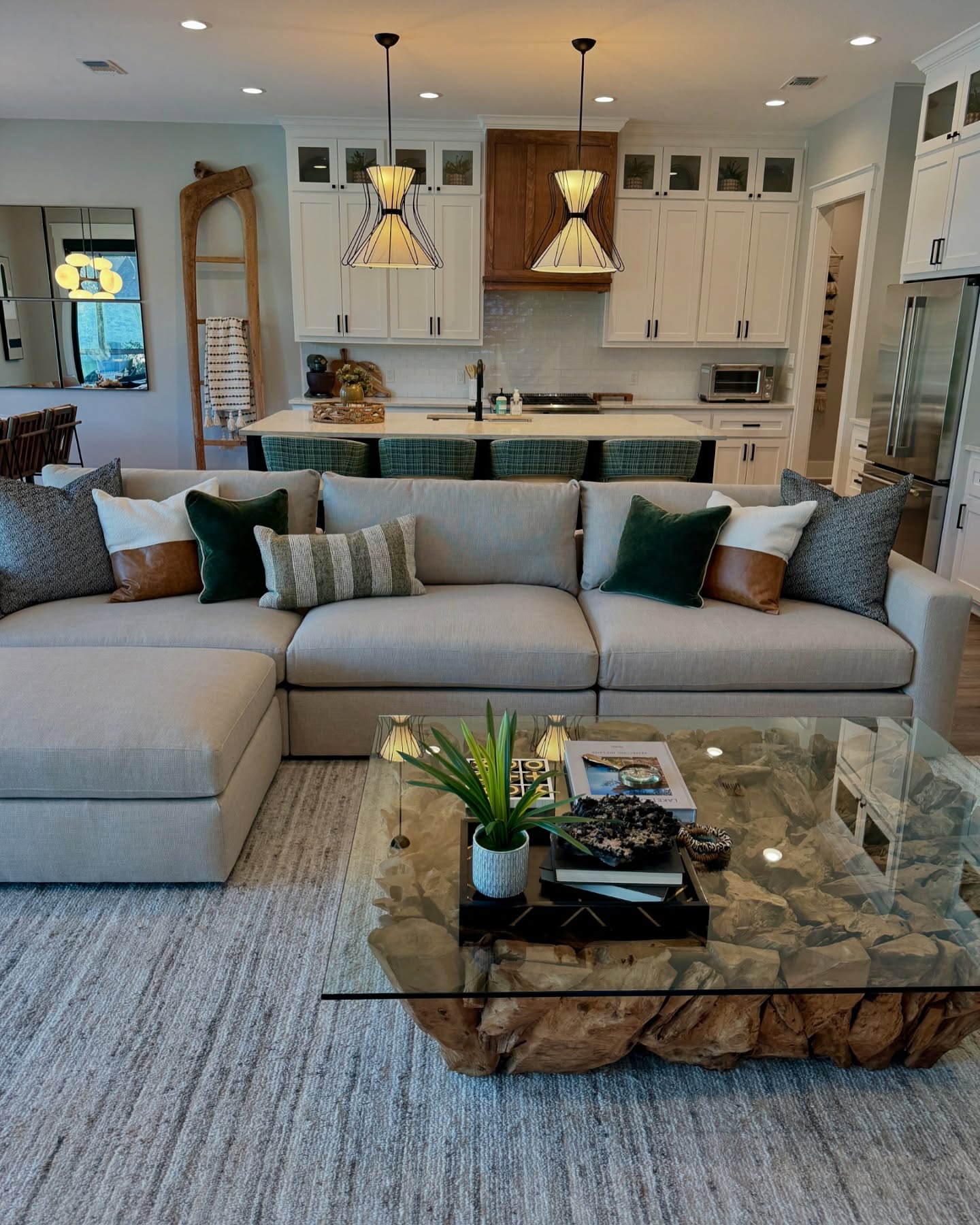 Open plan modern farmhouse living room with cream sectional sofa, leather and green pillows, and a driftwood and glass coffee table facing a white kitchen with green barstools