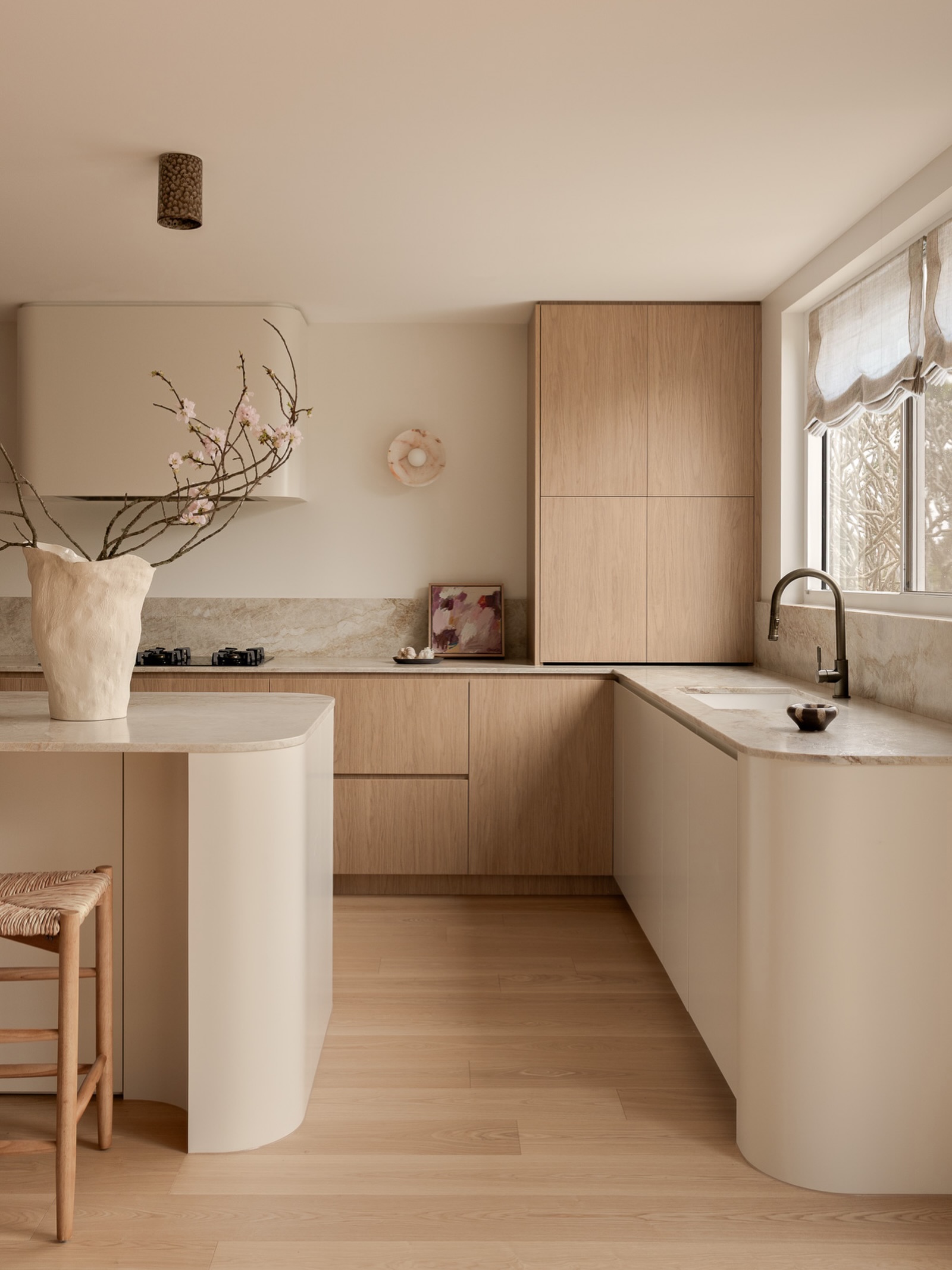 Minimal curved kitchen with a tall light oak flat-panel cabinet, a cream curved plaster island, travertine counters and backsplash, cherry blossom branches, and a woven rush stool