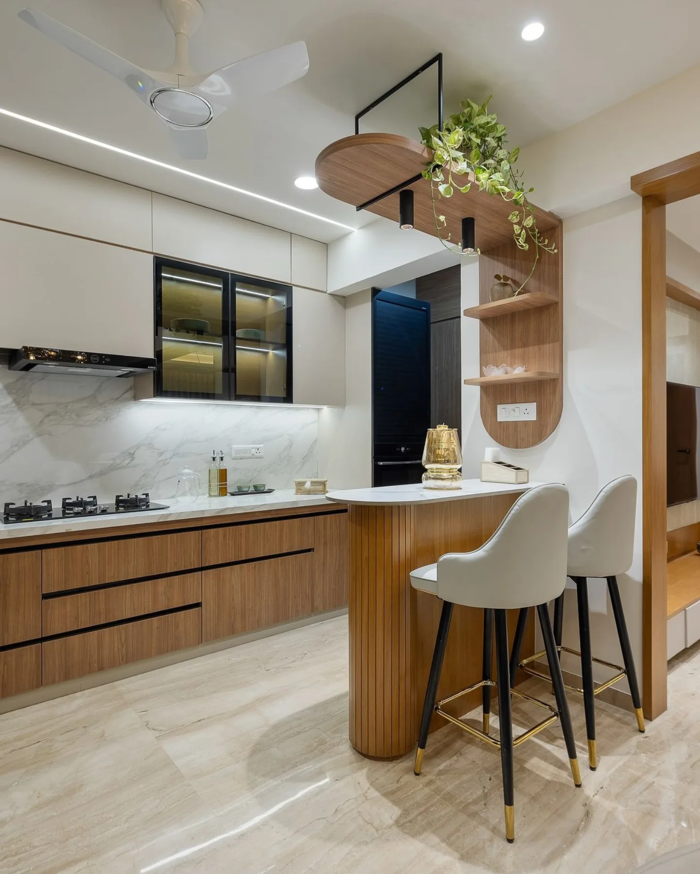 Modern kitchen with wood grain lower cabinets, white marble backsplash and counter, a curved peninsula wrapped in vertical wood slat paneling with a white marble top, two white and gold upholstered bar stools, and a round wood wall shelf with trailing pothos greenery