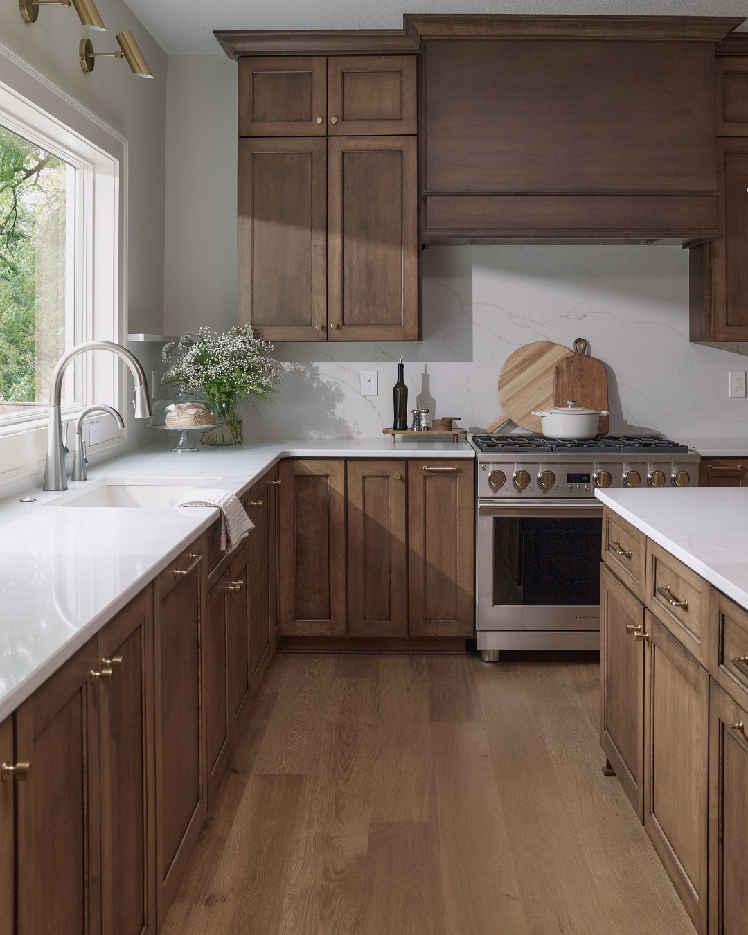 Medium stained walnut shaker kitchen cabinets with a soft white marble backsplash, brass wall sconces above the sink and a wood matching range hood