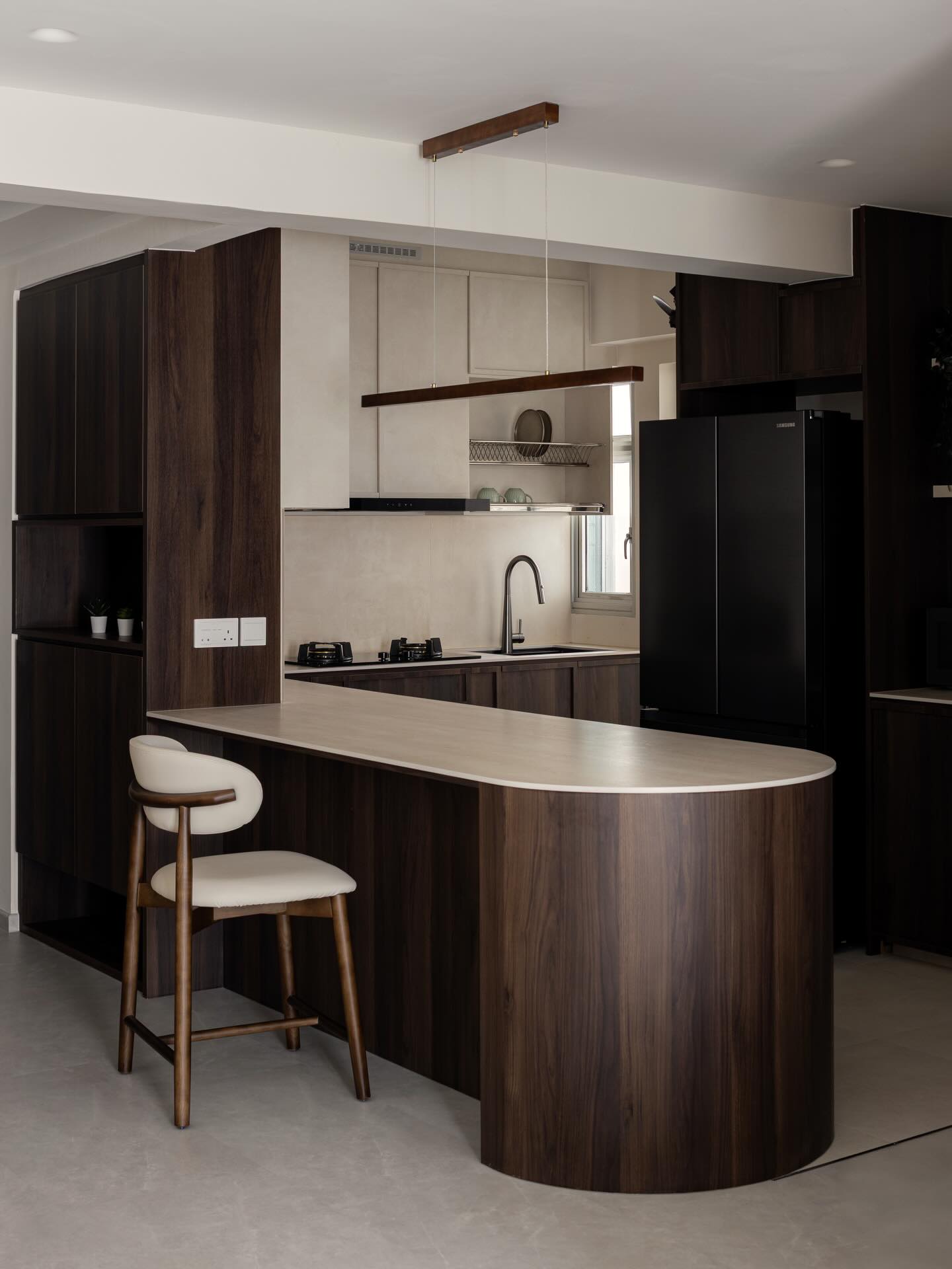 Dark walnut rounded peninsula with a pale stone countertop, a single cream and wood bar stool, a black Samsung fridge, and a beige stone backsplash behind
