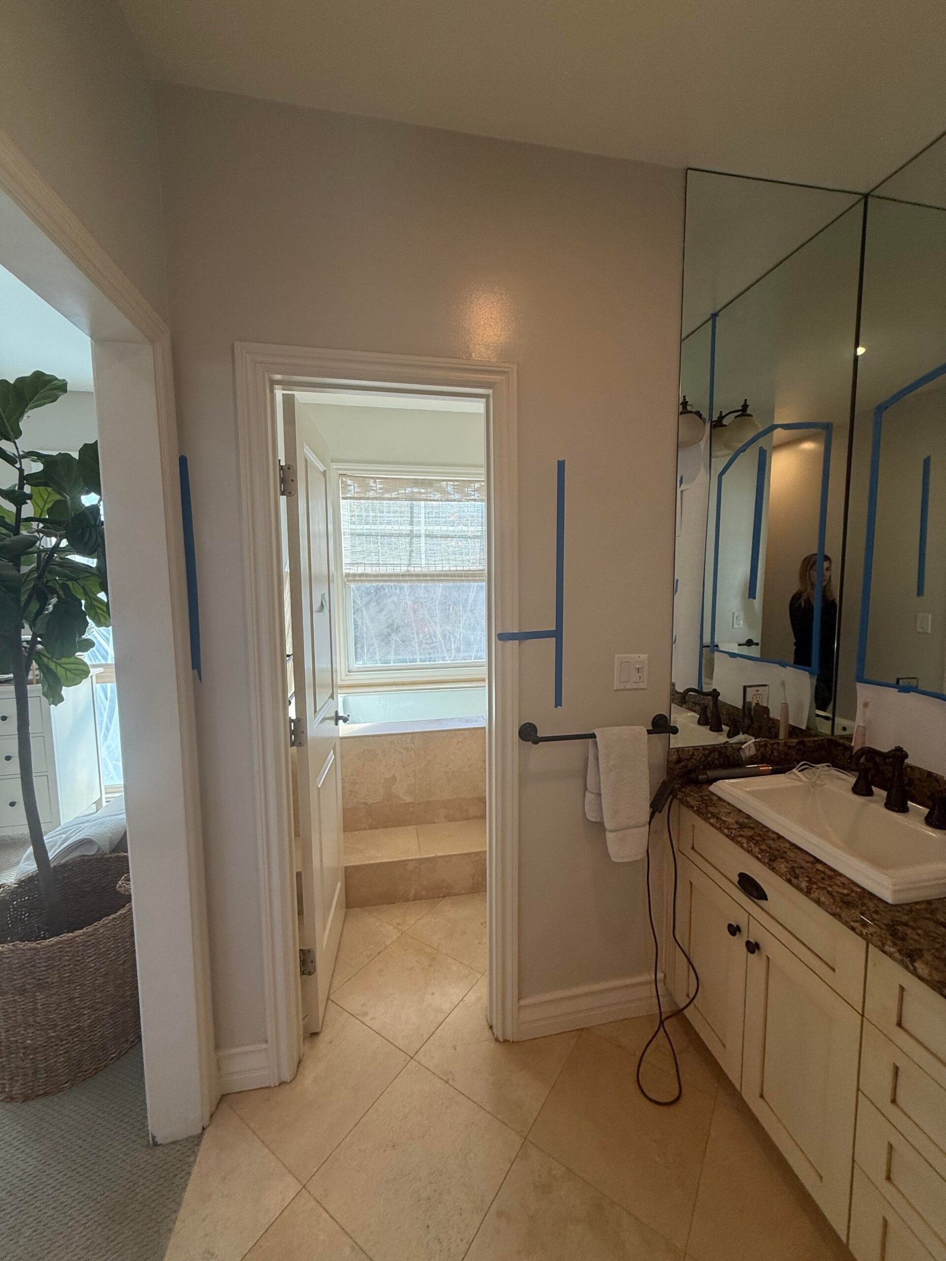 View through a cream wood door into a dated travertine tub and shower room, with another cream vanity and sheet mirror visible to the right.