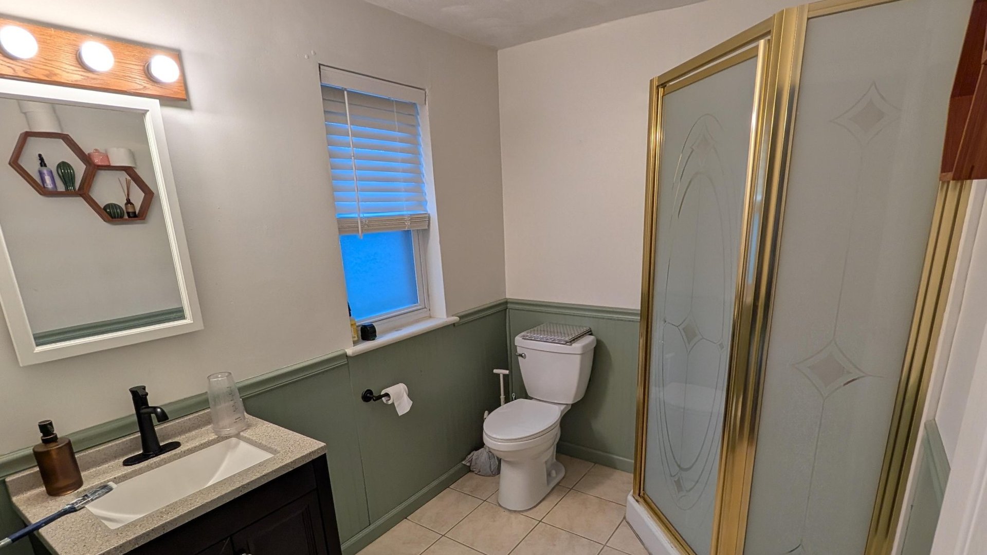Dated small bathroom with sage green painted wainscoting and off-white upper walls, a dark wood vanity with speckled laminate top and black faucet, an oak-trim Hollywood vanity light with three round bulbs above a plain mirror, a brass-framed corner shower stall with etched glass panels, and a white toilet below a small frosted window.