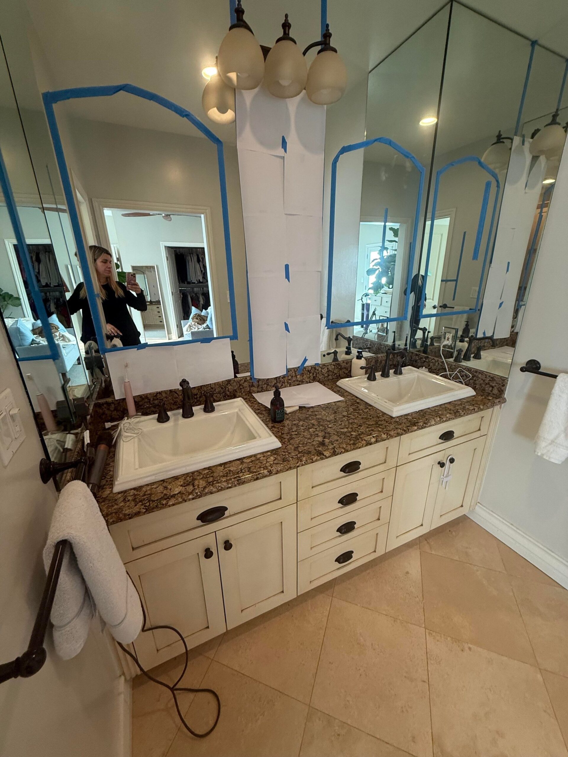 Dated primary bathroom vanity with cream raised-panel cabinets, speckled brown granite counters, drop-in sinks, oil-rubbed bronze pulls, and a wall-to-wall sheet mirror taped off with blue painter's tape.