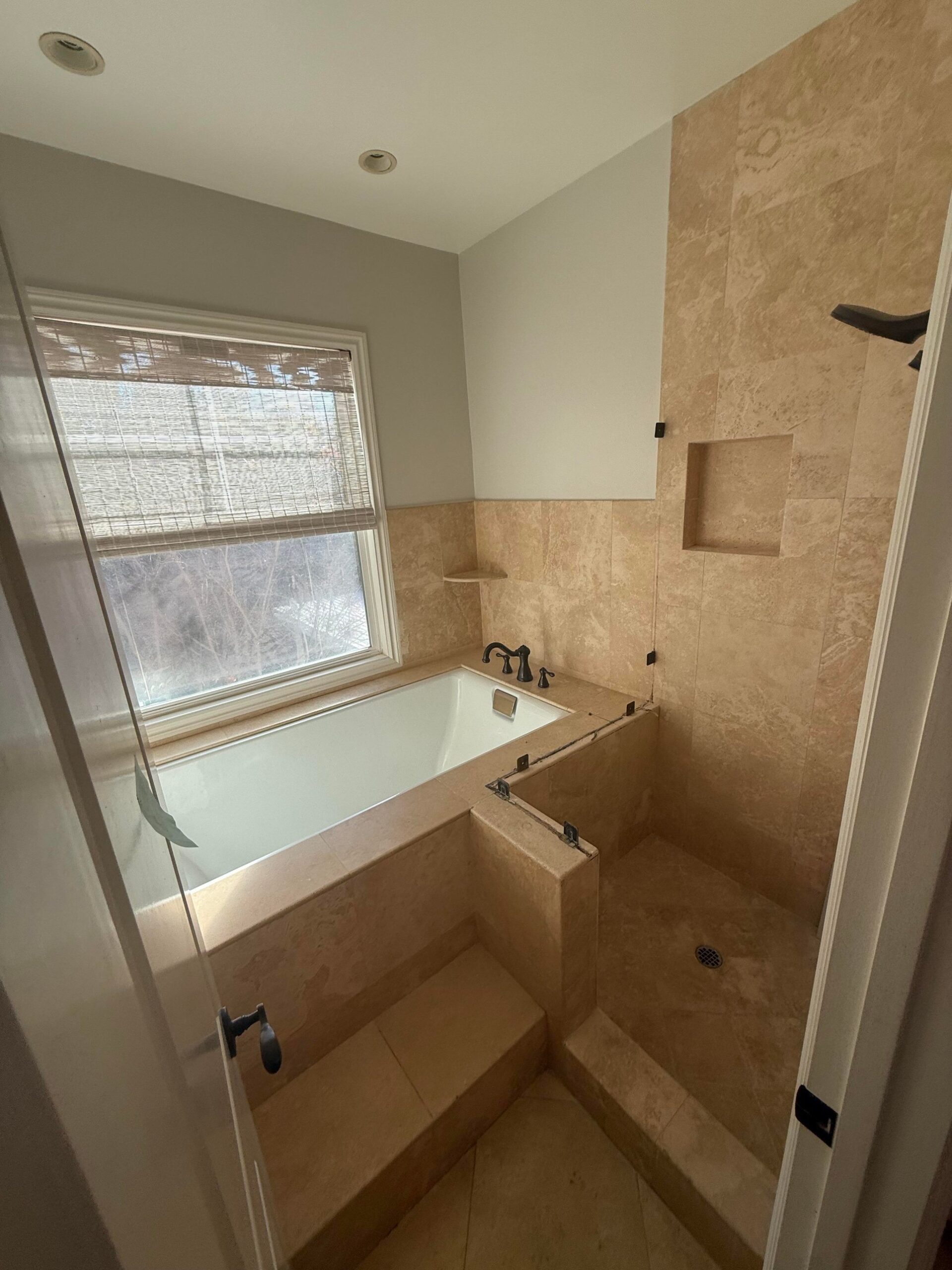 Dated travertine primary bath with a deck-mounted tub under a window, oil-rubbed bronze faucet, bamboo roller shade, and a tiled walk-in shower with no door, mid-remodel.