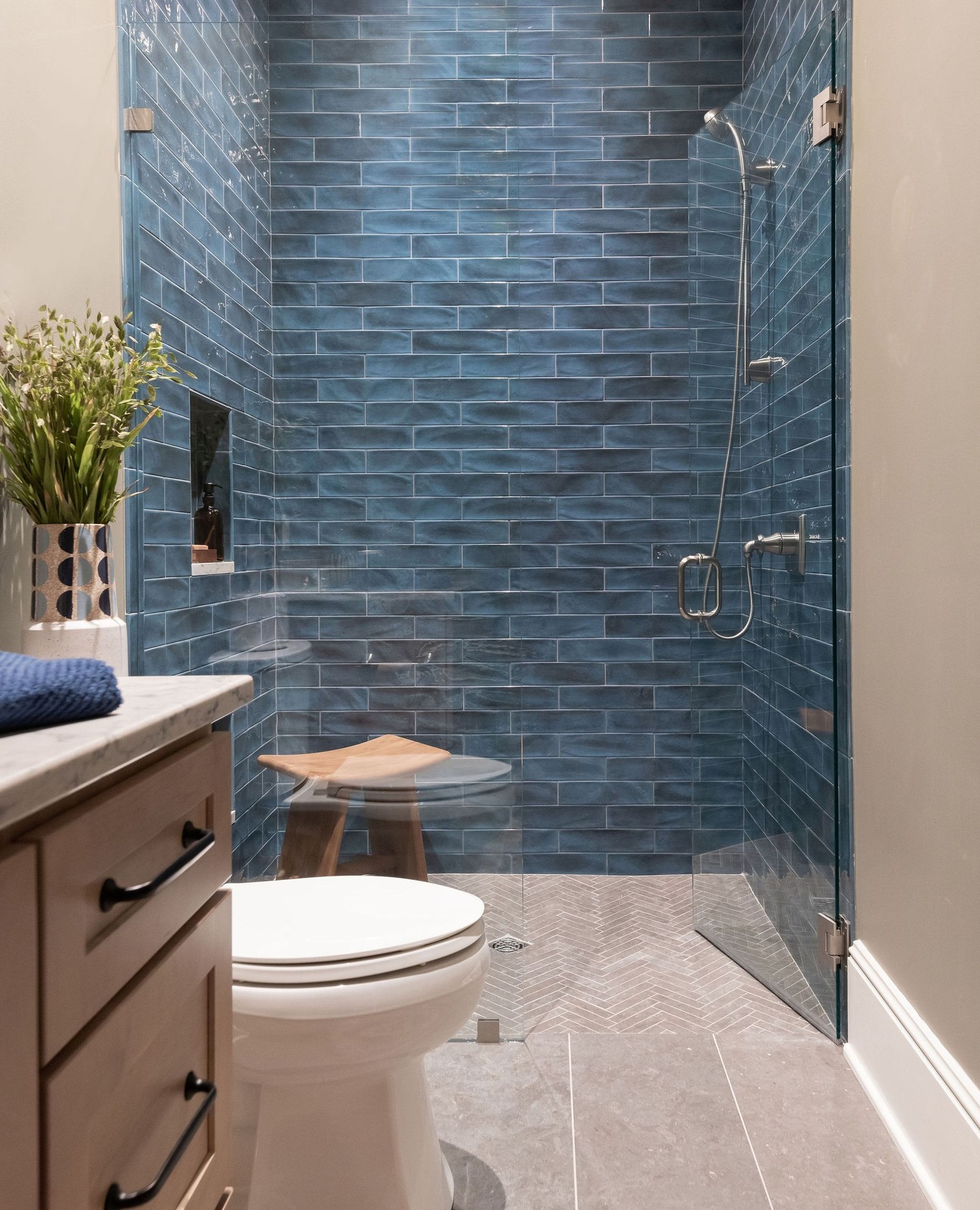Guest bathroom shower with glossy deep blue handmade subway tiles in a running bond pattern, a gray herringbone floor, a wood seat, a glass panel door, and a wood vanity with a marble top